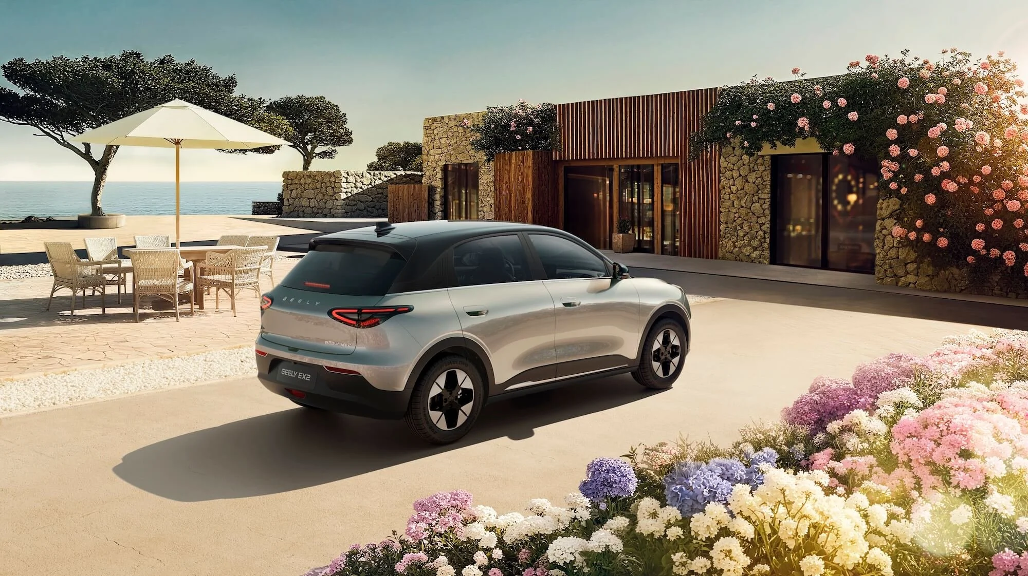 Coche eléctrico Geely EX3 estacionado en un entorno costero con jardines de flores y una casa moderna de piedra y madera, junto a una mesa con sombrilla bajo árboles y vista al mar en el fondo.
