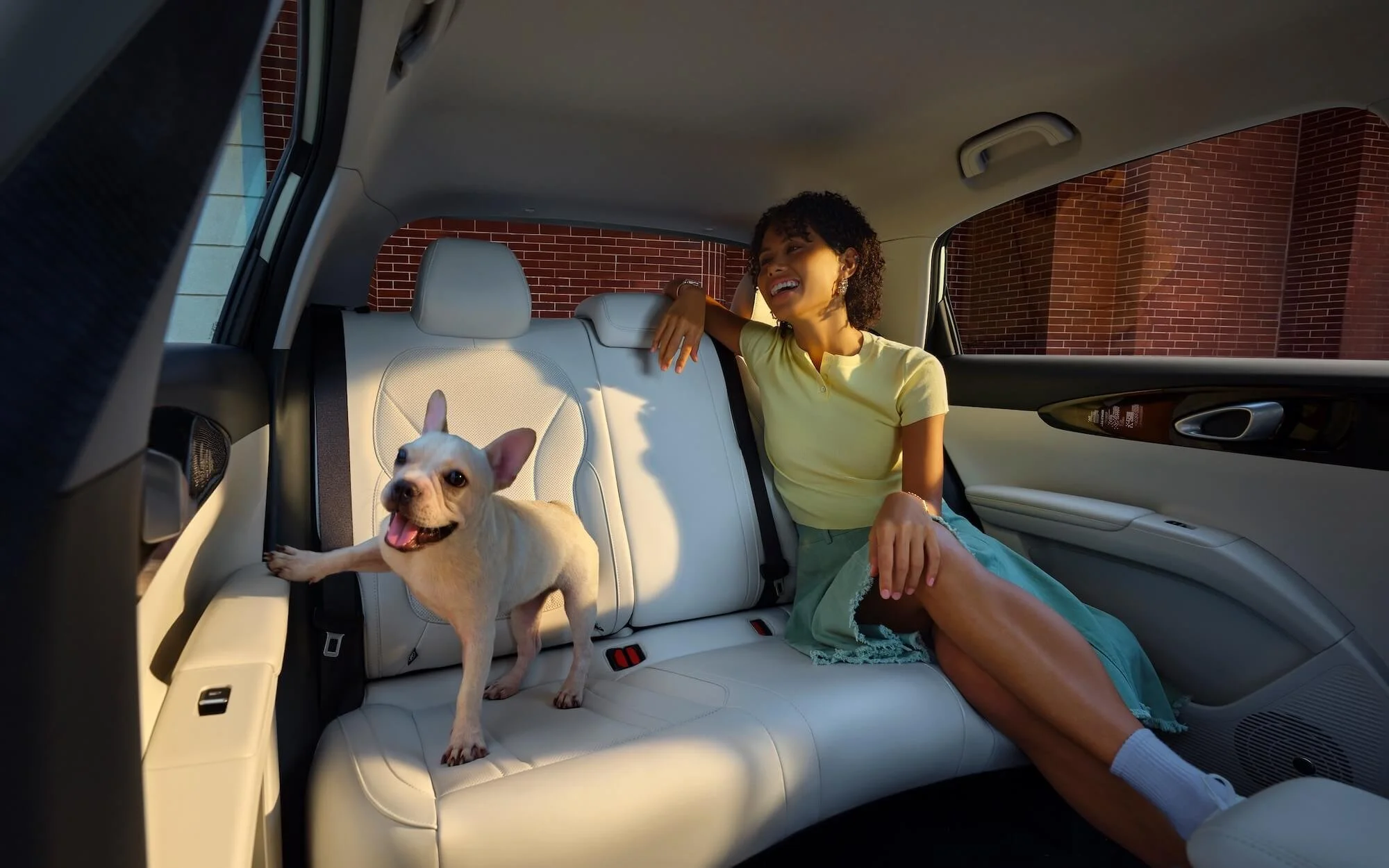 Una mujer sonriente sentada en la parte trasera de un coche con un perro pequeño en el asiento junto a ella.
