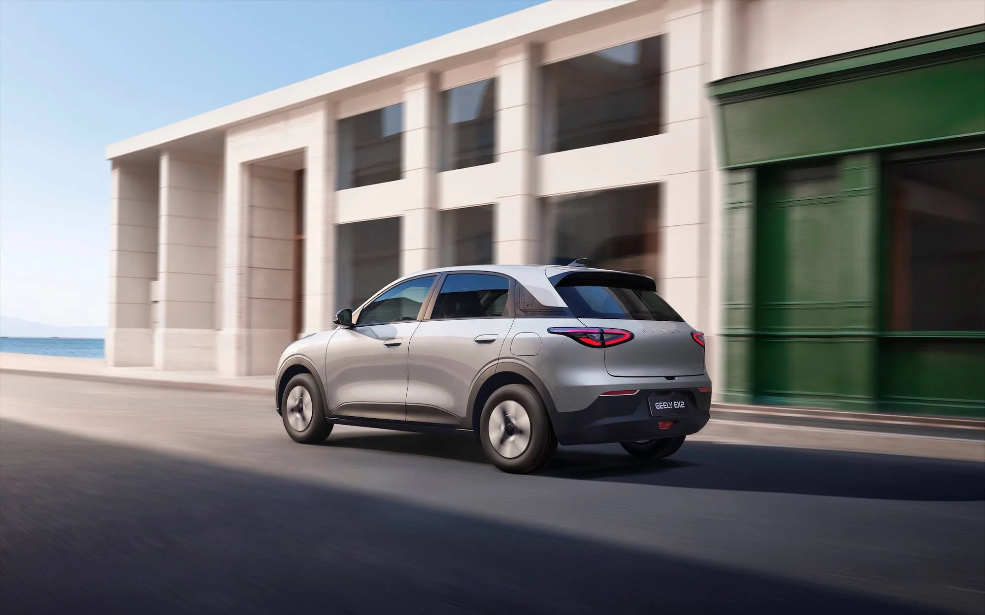 Auto eléctrico gris conduciendo por una calle urbana con edificios modernos y cielo claro.
