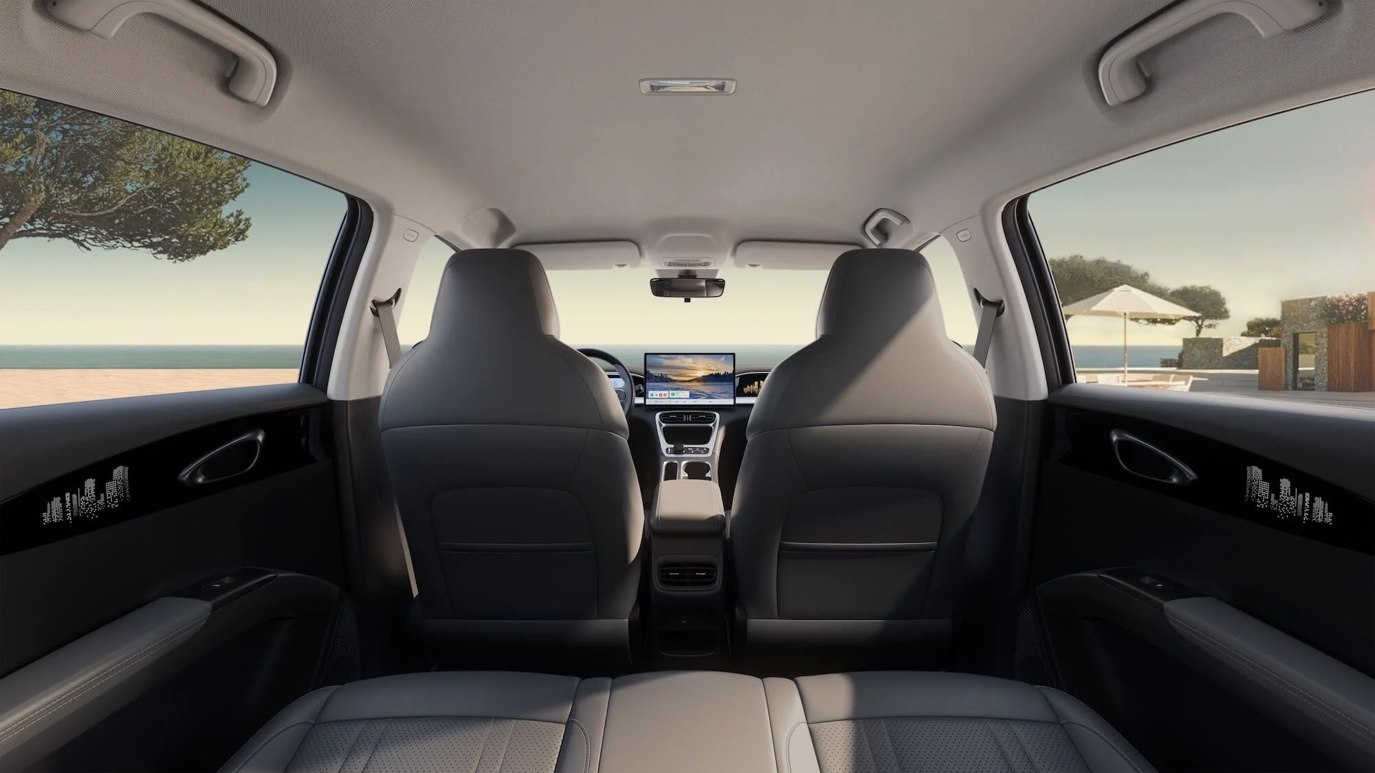 Interior de un vehículo con vista a la playa, con asientos de color gris y pantalla digital en el tablero, ventanas grandes con vista al mar y un área con sombrilla y muebles en el exterior.