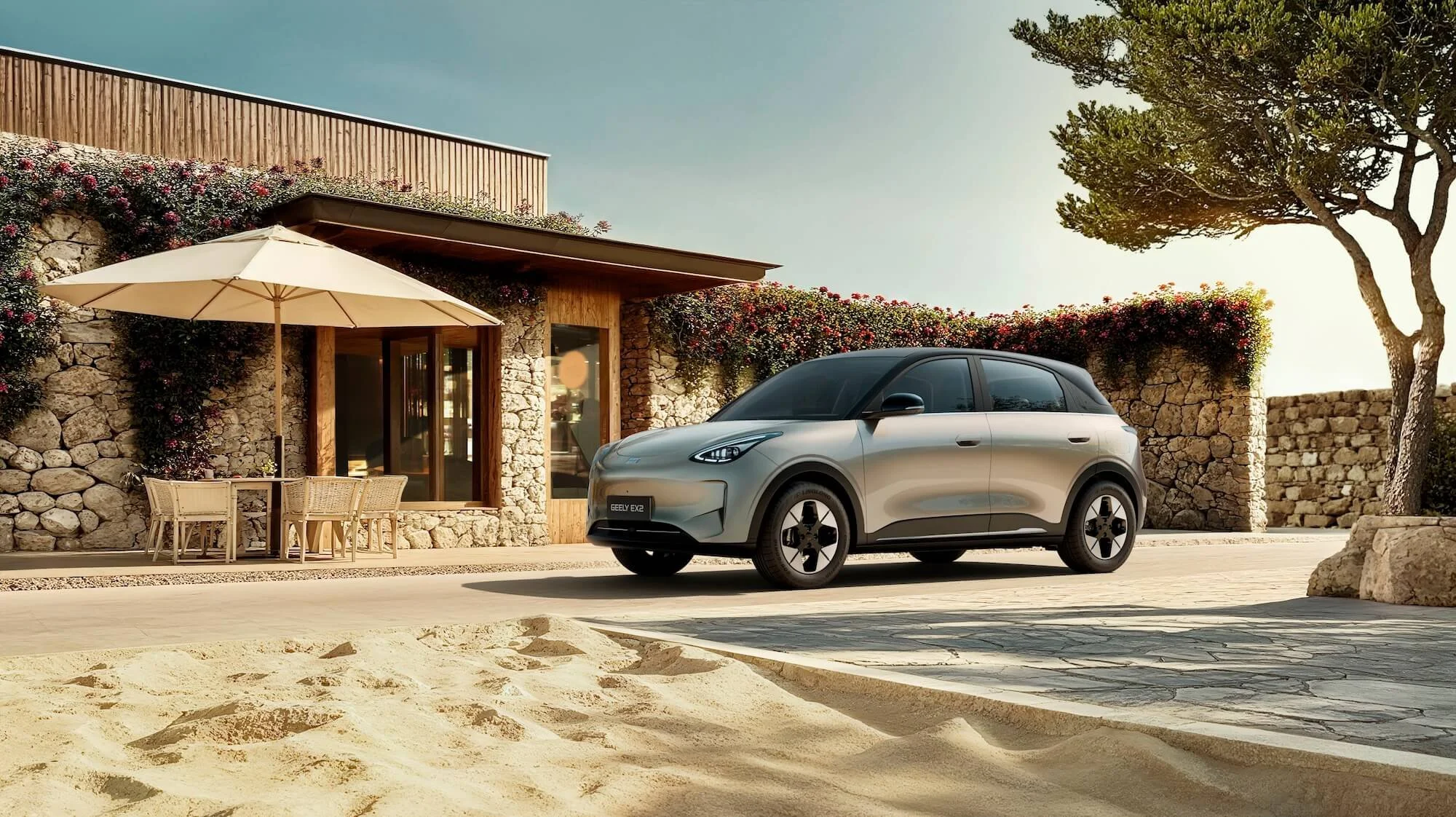 Coche eléctrico plateado estacionado frente a una casa de piedra con terraza y sombrilla, en un entorno soleado con árboles y vegetación.