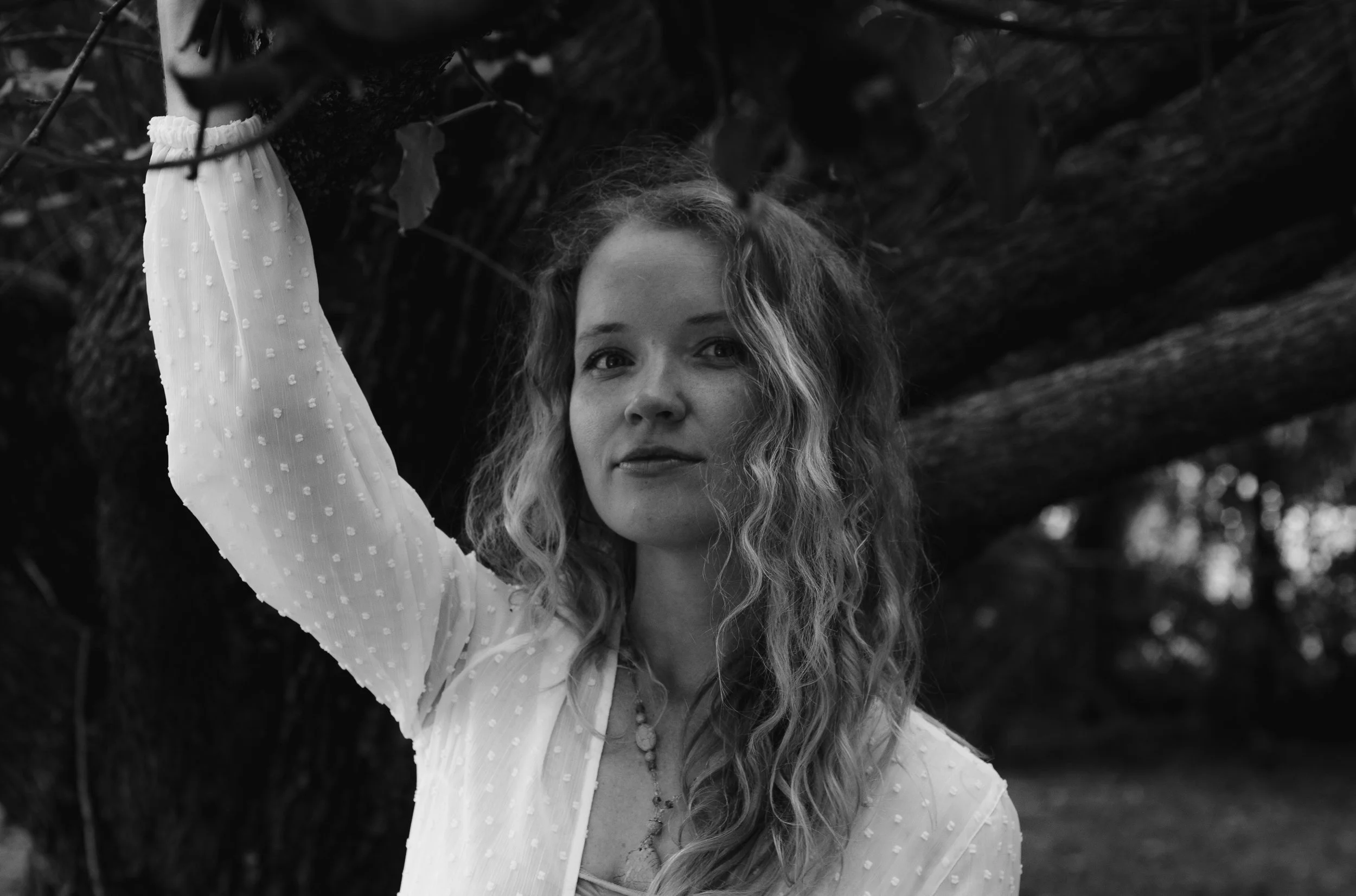 A woman with long, curly hair standing outdoors next to a large tree, wearing a light-colored, textured blouse with a necklace, looking at the camera.