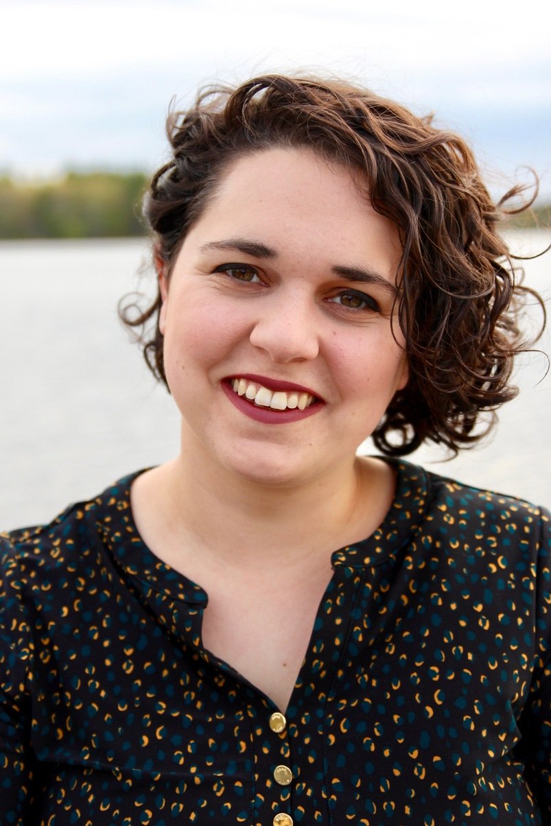 A woman with short, curly brown hair smiling outdoors near a body of water with a cloudy sky.