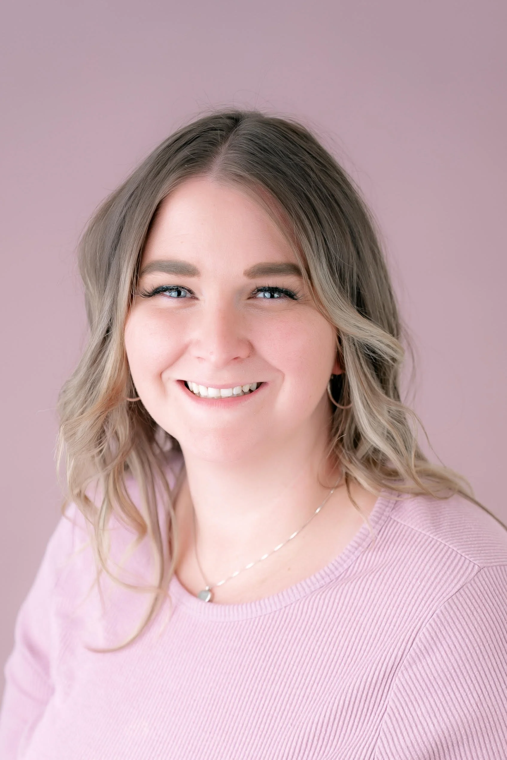 A woman with blonde, wavy hair smiling against a light pink background, wearing a pink top and jewelry.