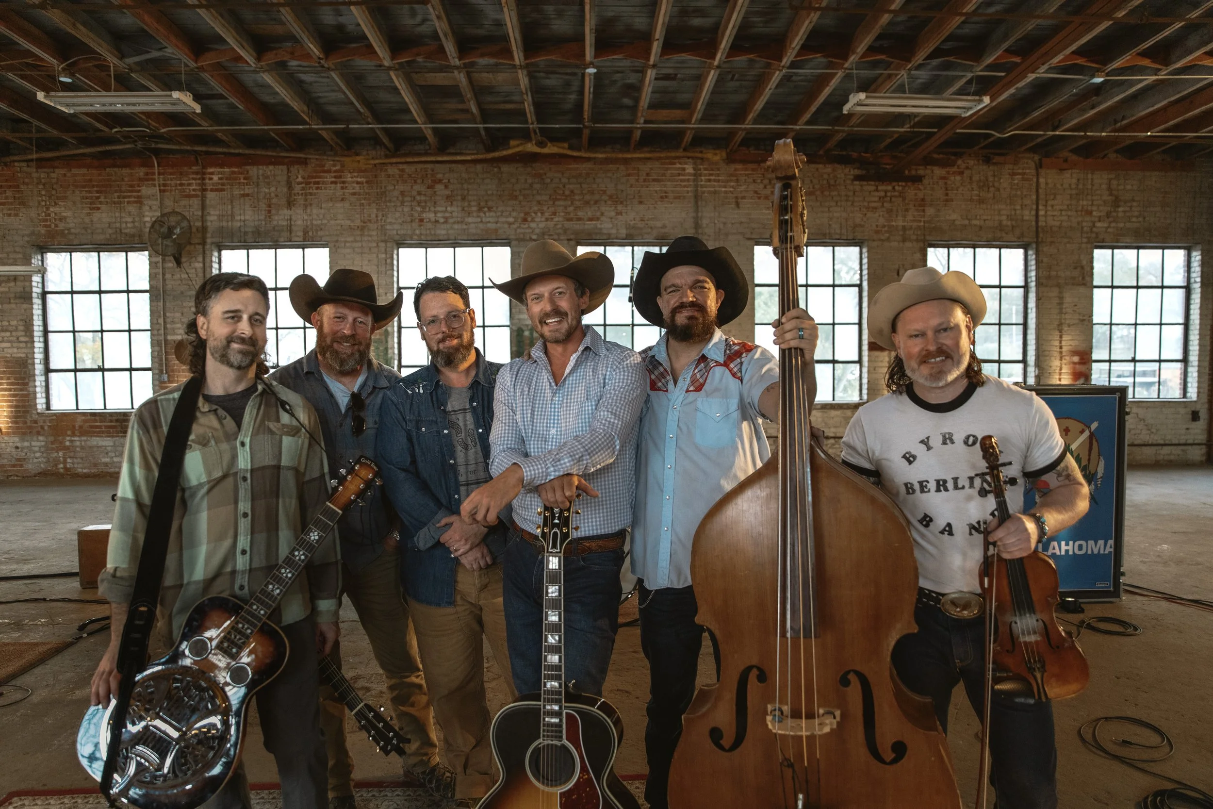 Six men holding musical instruments in an industrial-style room with brick walls and large windows, all wearing cowboy hats and casual Western-style clothing.