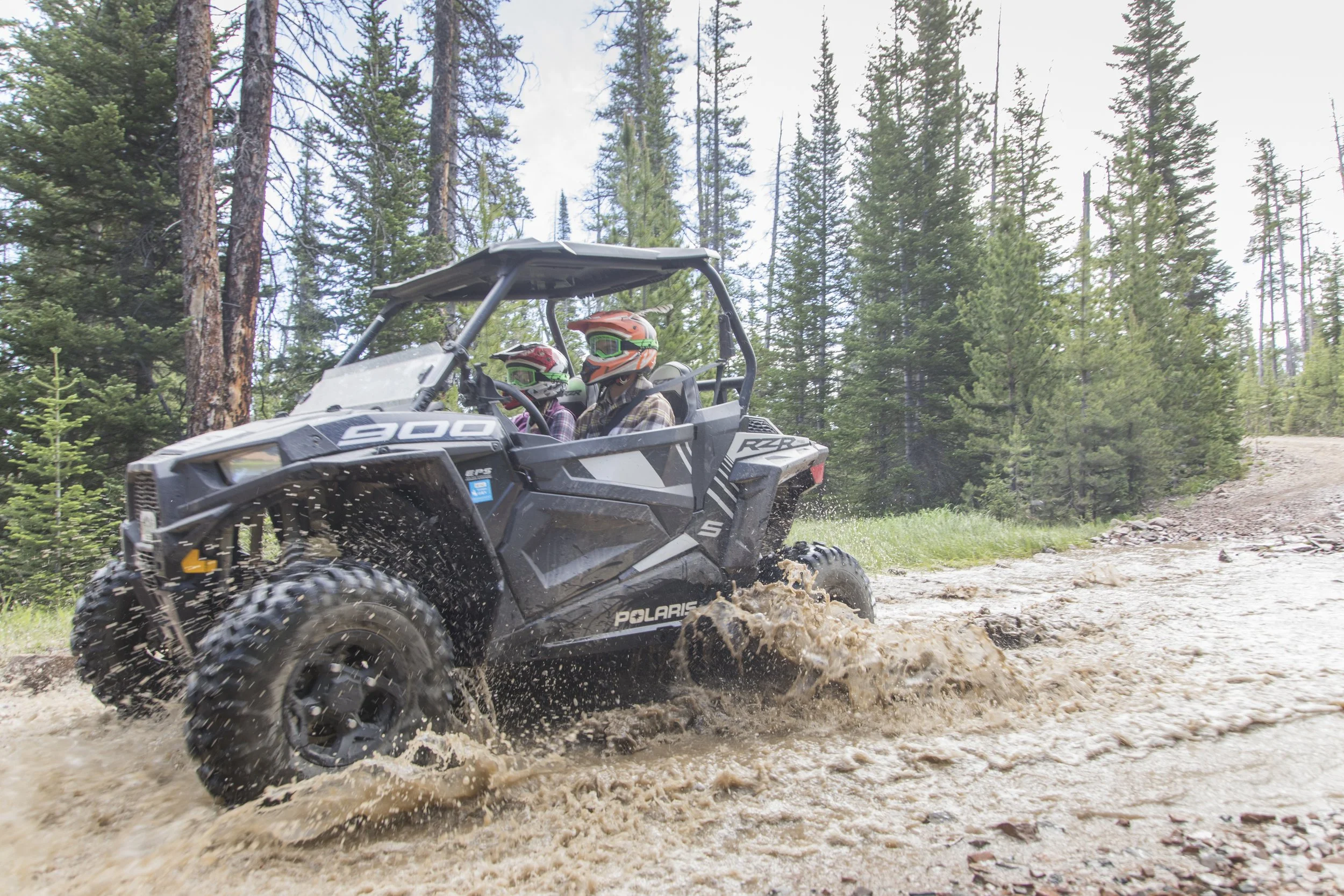 A Polaris RZR off-road vehicle driving through a dirt trail in a forest, with two people wearing helmets inside.
