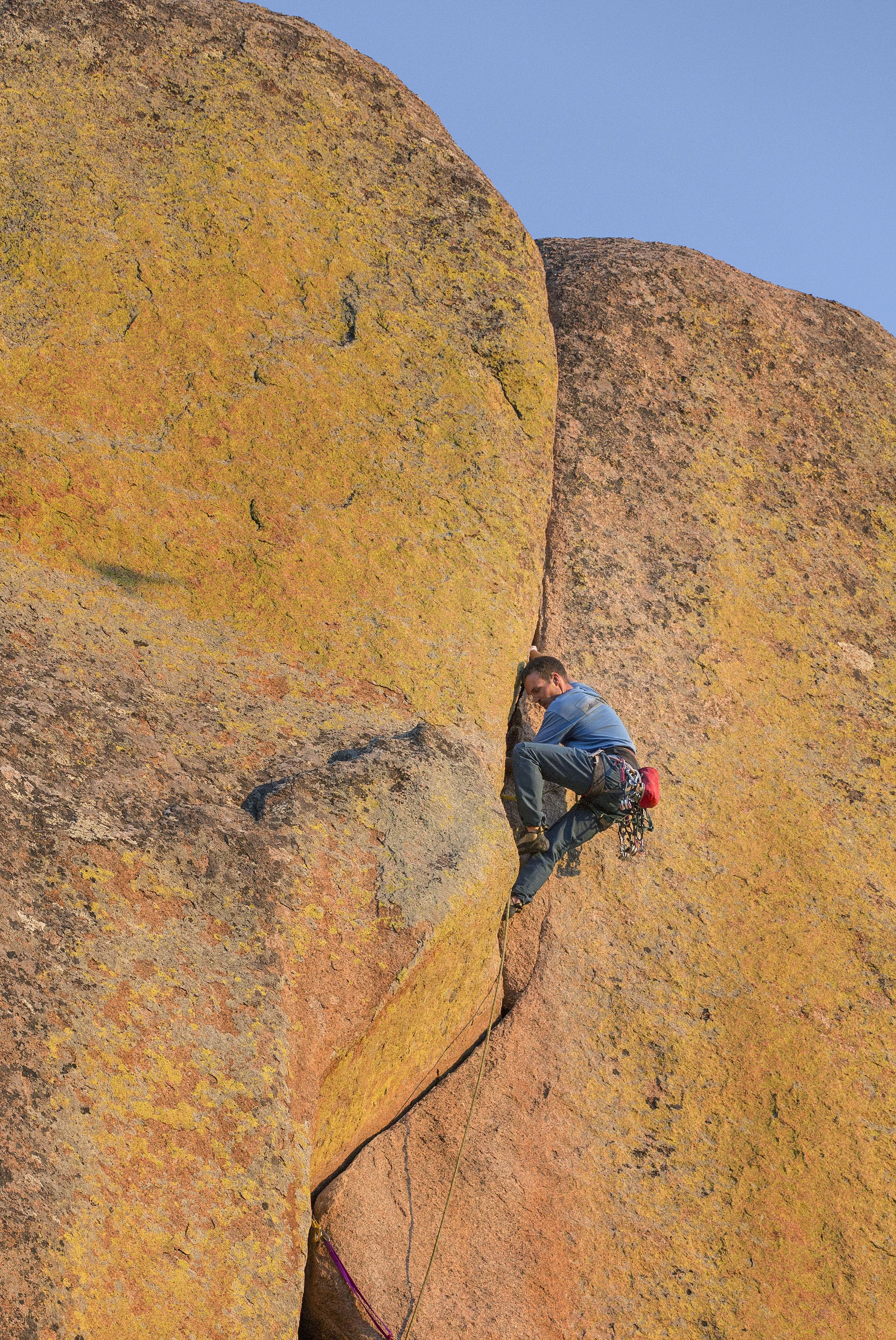 A man in climbing gear is rock climbing on a large, orange-tinted granite boulder under a clear blue sky.
