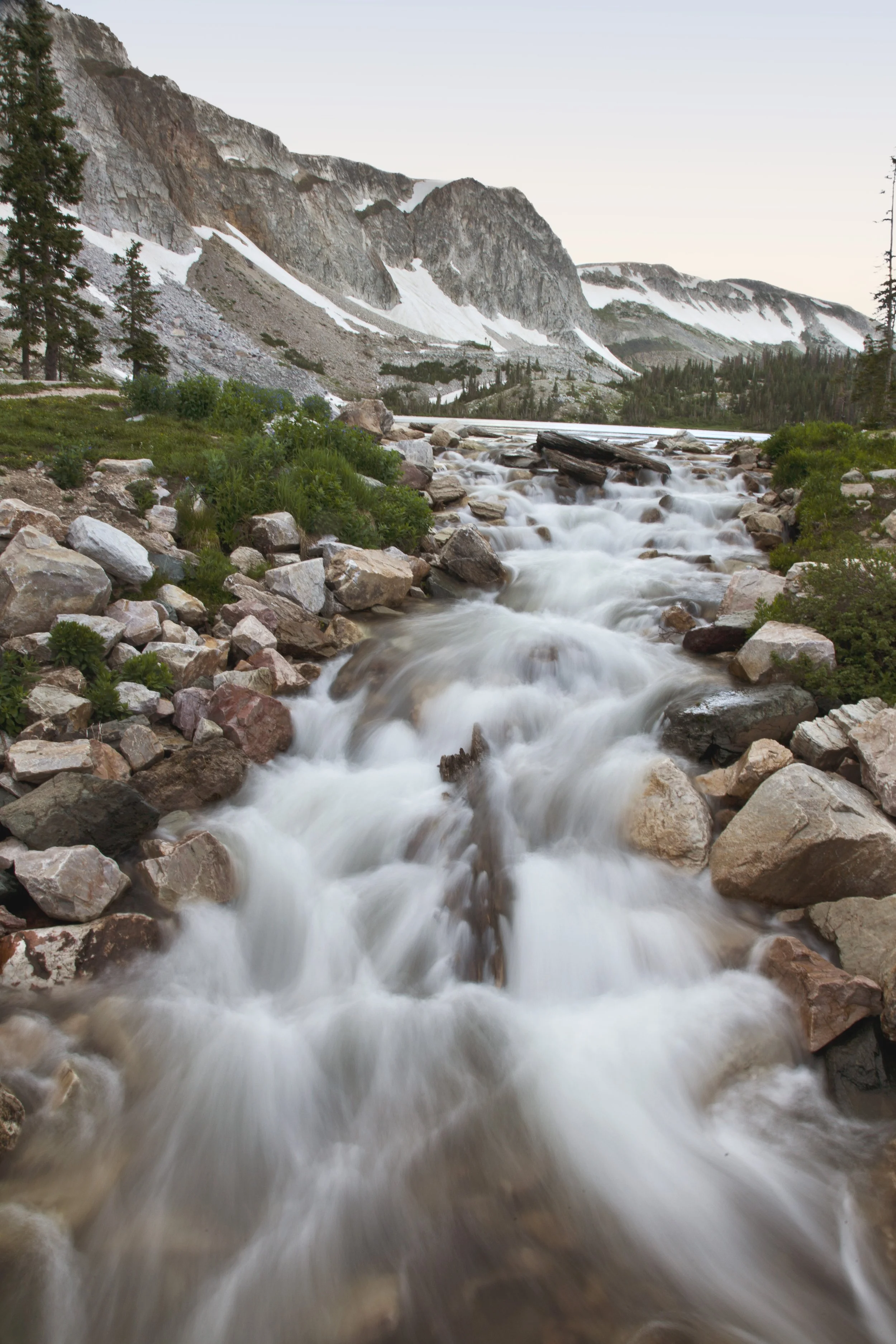 Flowing mountain river with rocks and greenery, snow-capped mountains in the background, and trees along the river bank.
