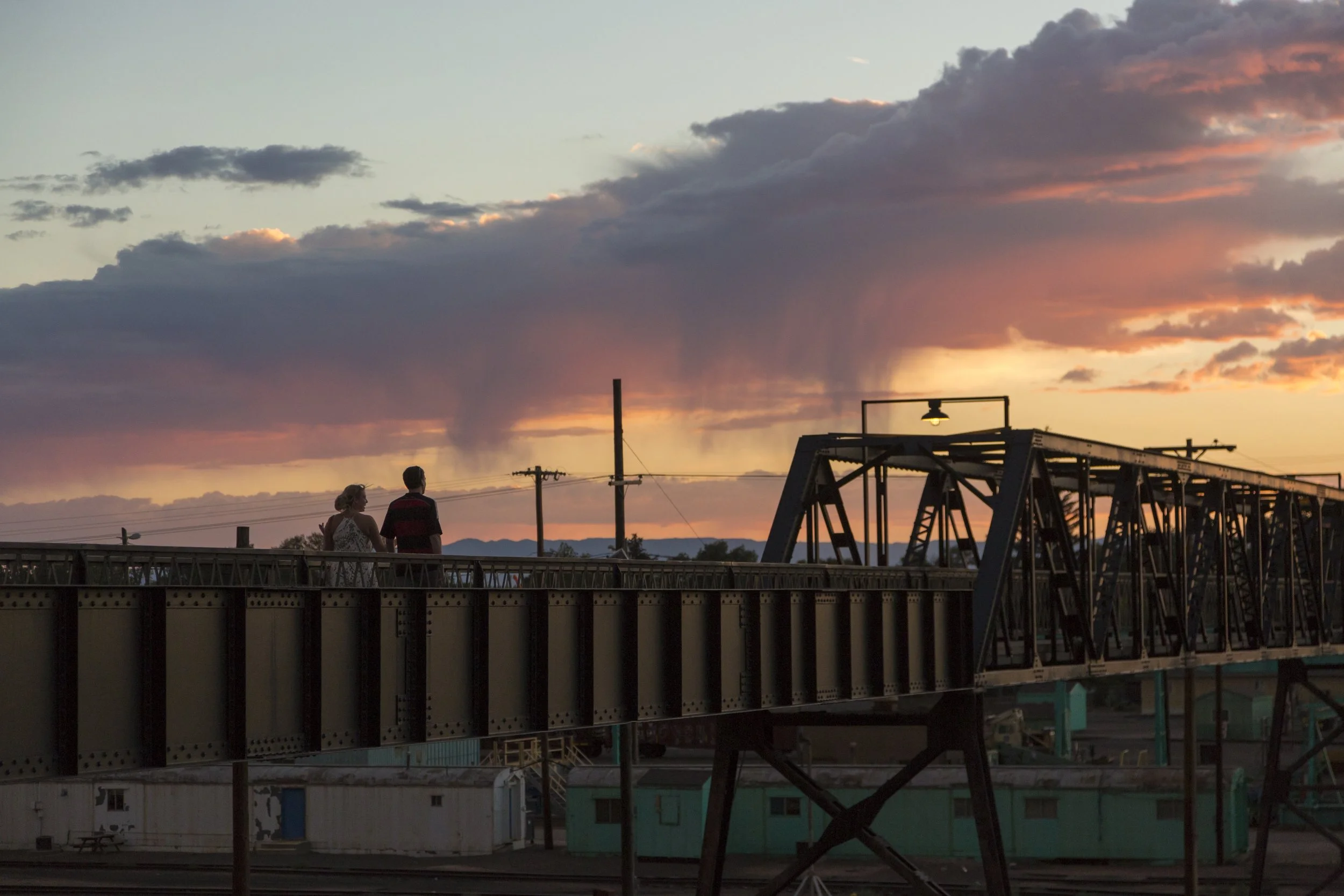 Two people walking on a bridge at sunset with a dramatic sky and clouds, industrial structures in the foreground, and mountains in the distance.