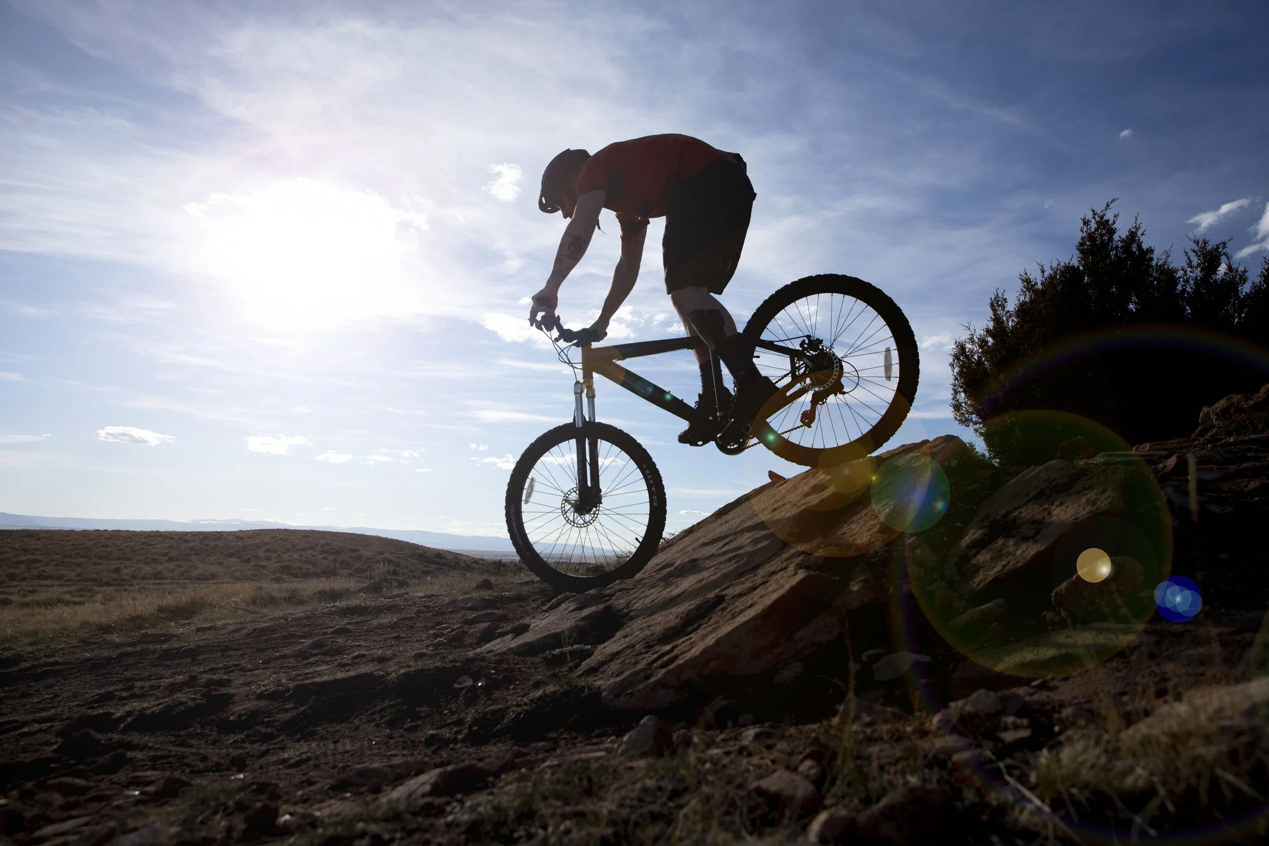 Bicycle rider in mid-air on a rocky trail with the sun shining behind, lens flare visible.