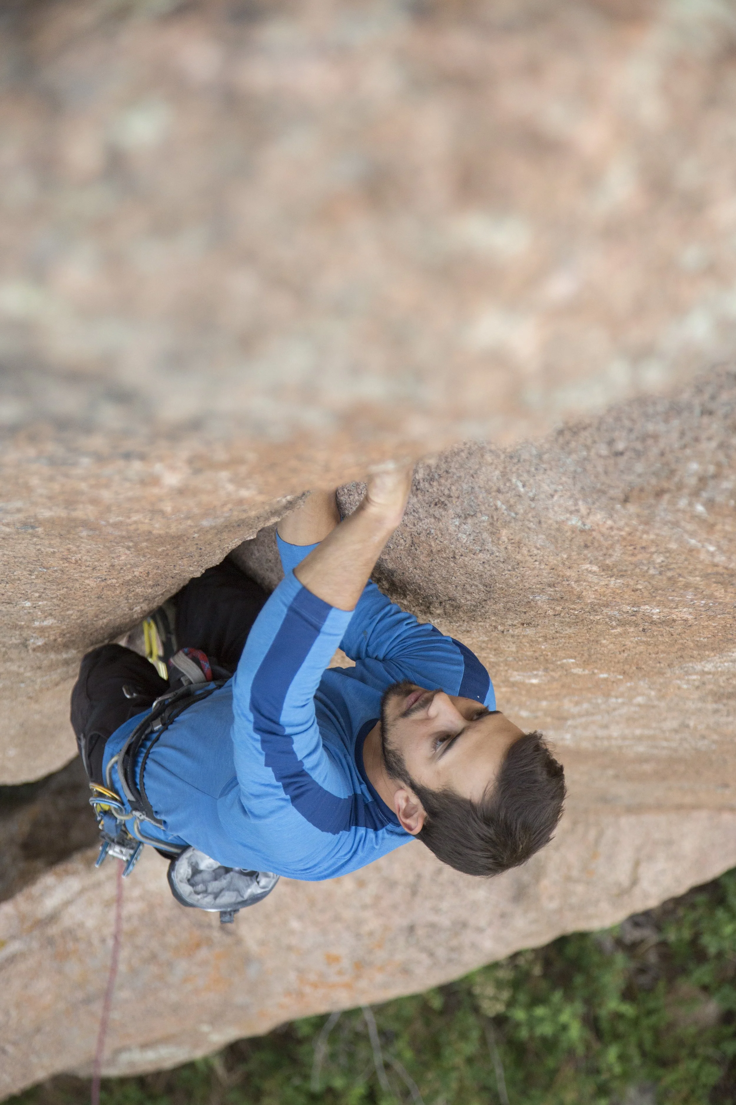 A man rock climbing on a reddish-brown cliff, wearing a blue long-sleeve shirt and climbing gear.