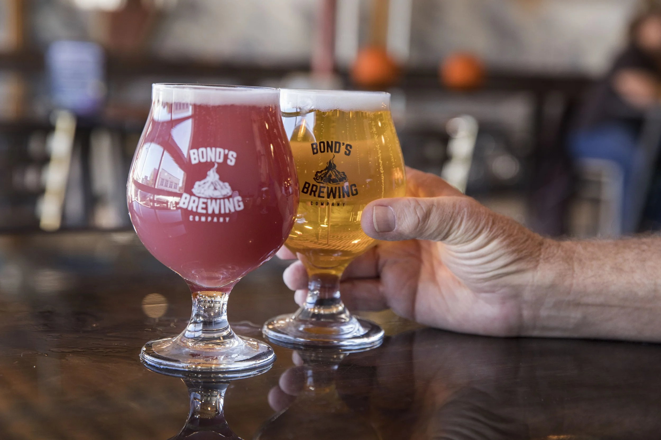 Two beer glasses with the logo 'Bond's Brewing' on a dark polished table, one containing pink/red beer and the other containing golden beer, with a hand holding the golden beer glass in the background of a rustic bar or brewery setting.