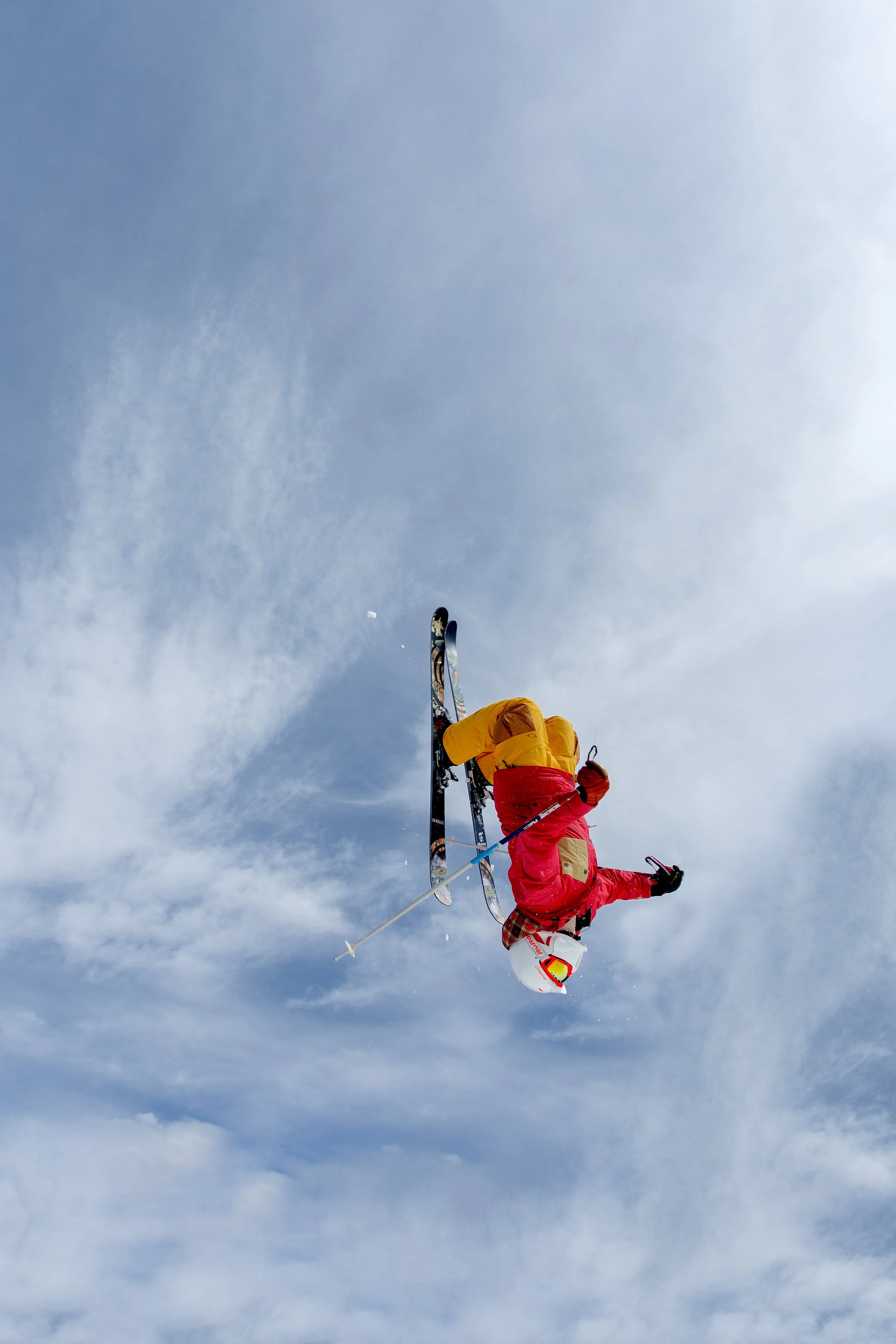 A person dressed in red and yellow winter gear is mid-air performing a trick on skis against a partly cloudy sky.