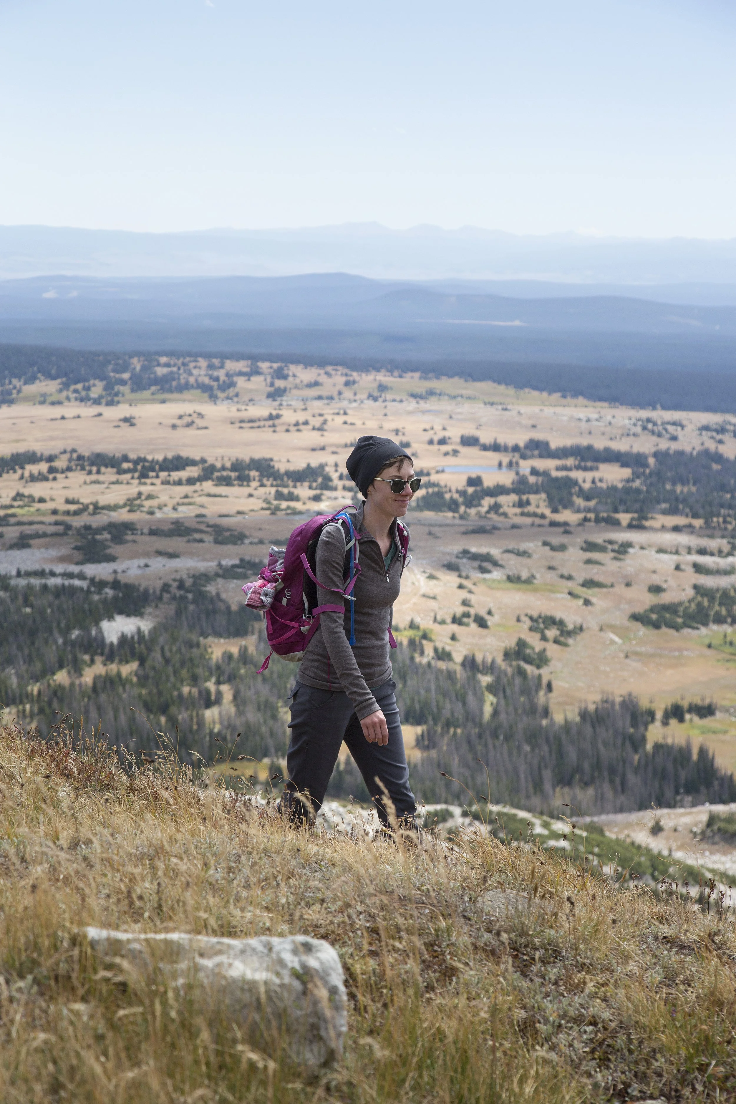 A woman hiking in a mountainous landscape with a valley below, wearing sunglasses, a beanie, and a purple backpack.