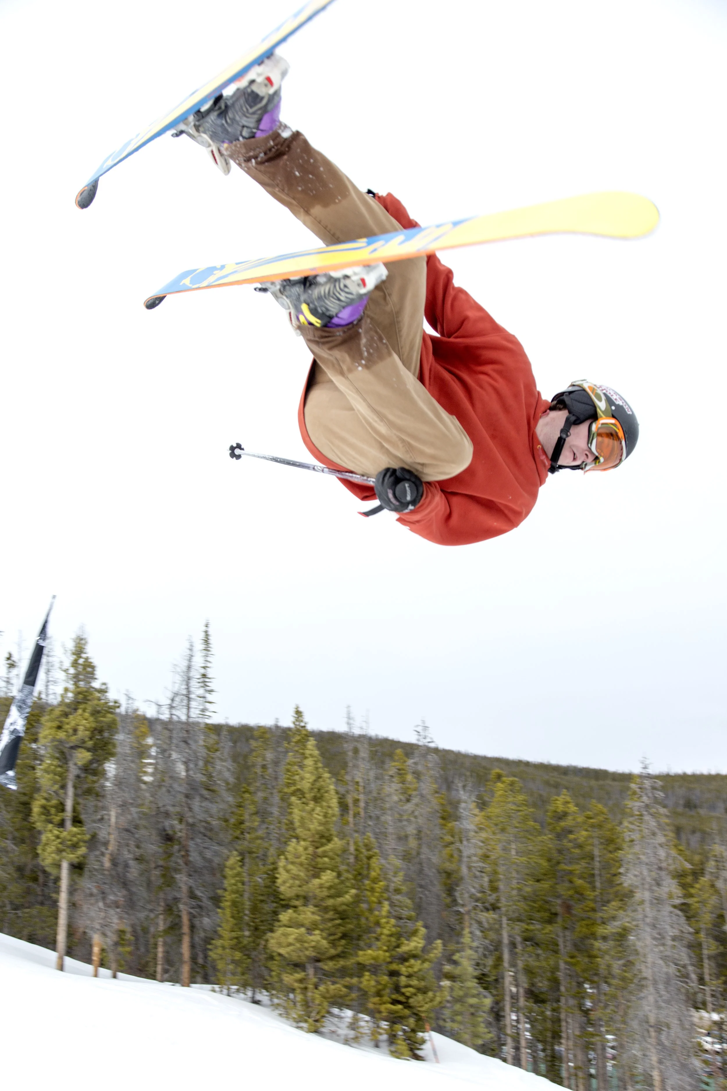 A skier wearing a red jacket, tan pants, purple gloves, and a helmet with goggles is mid-air performing a stunt on a snowy slope during daytime with a forested hill in the background.