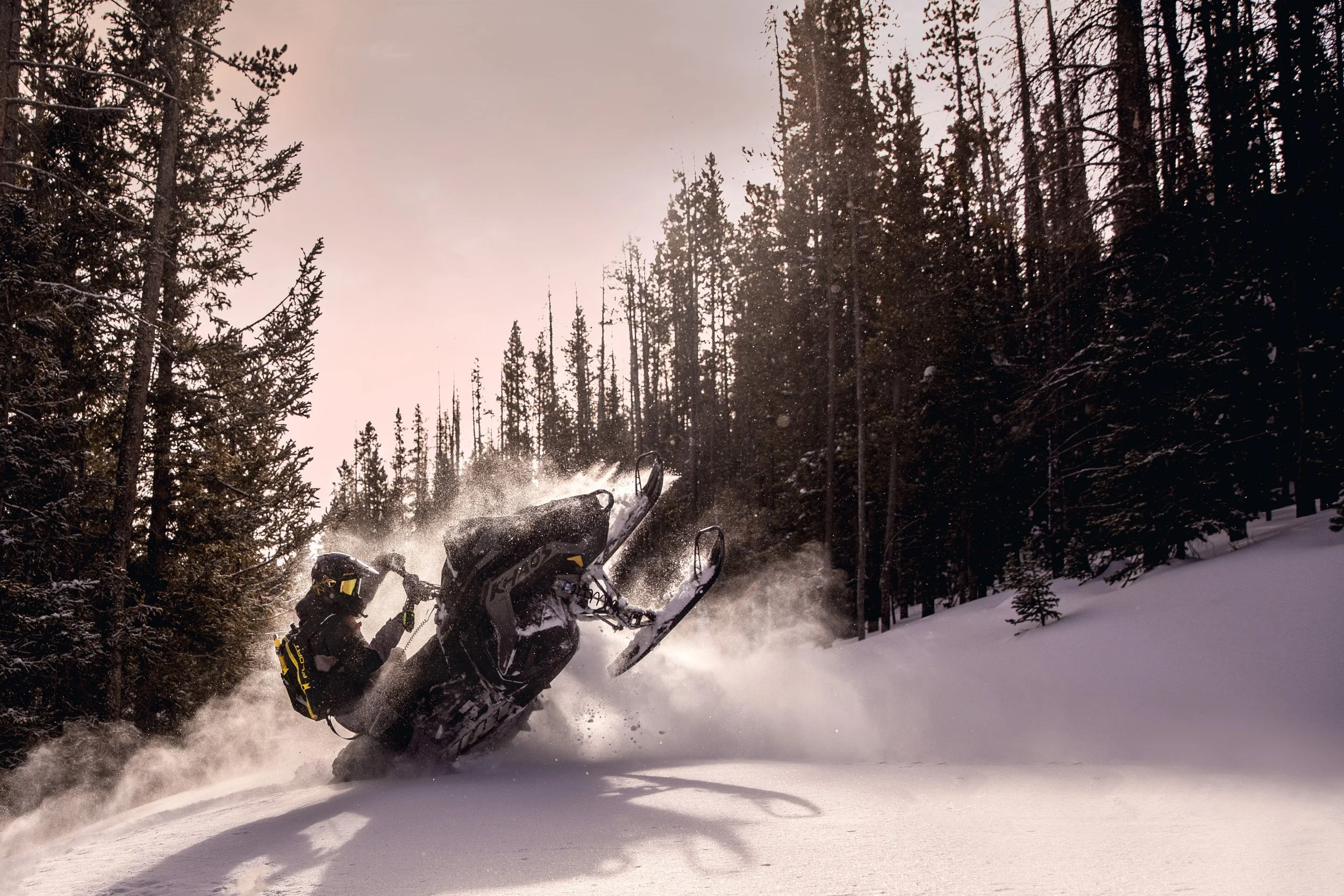 A person riding a snowmobile in a snowy forest, kicking up snow as they navigate through trees during sunset.