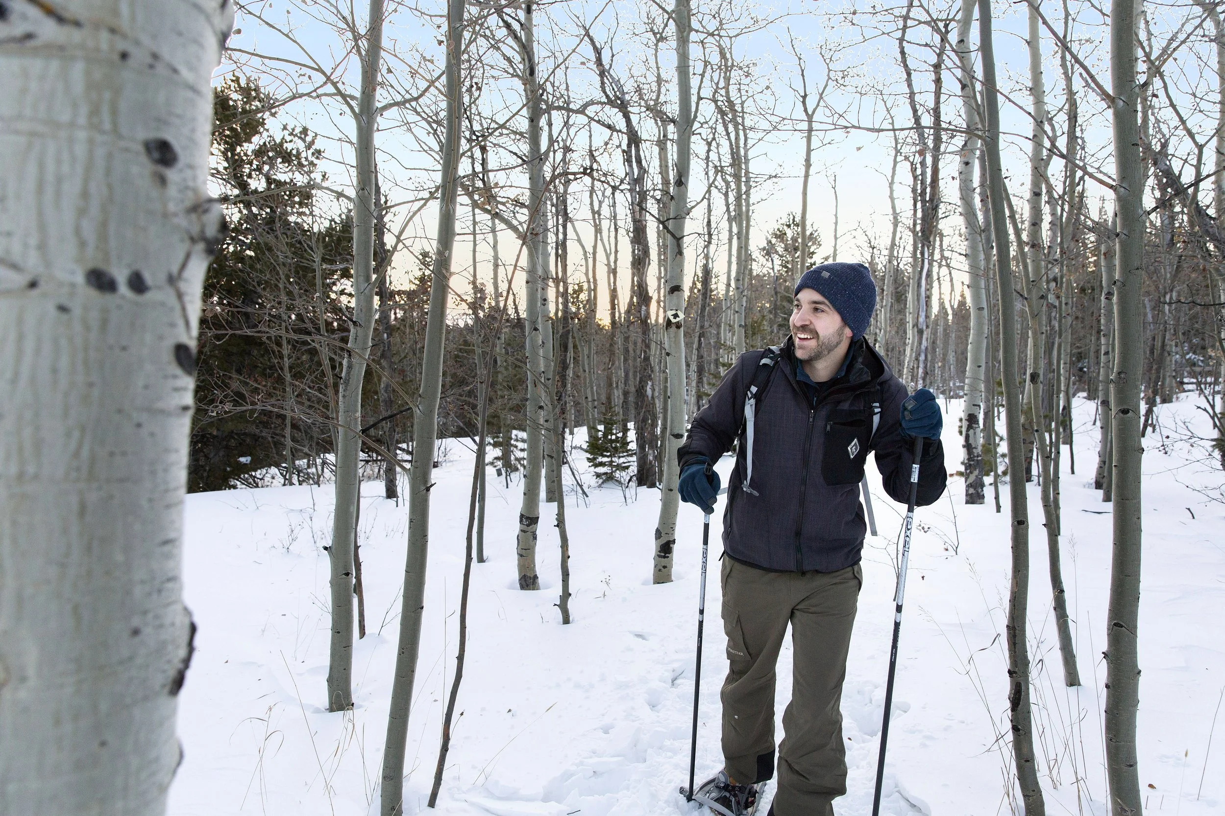 A man snowshoe hiking through a snowy forest during winter, wearing a beanie, gloves, and a black jacket, holding snowshoe poles, with leafless trees around.