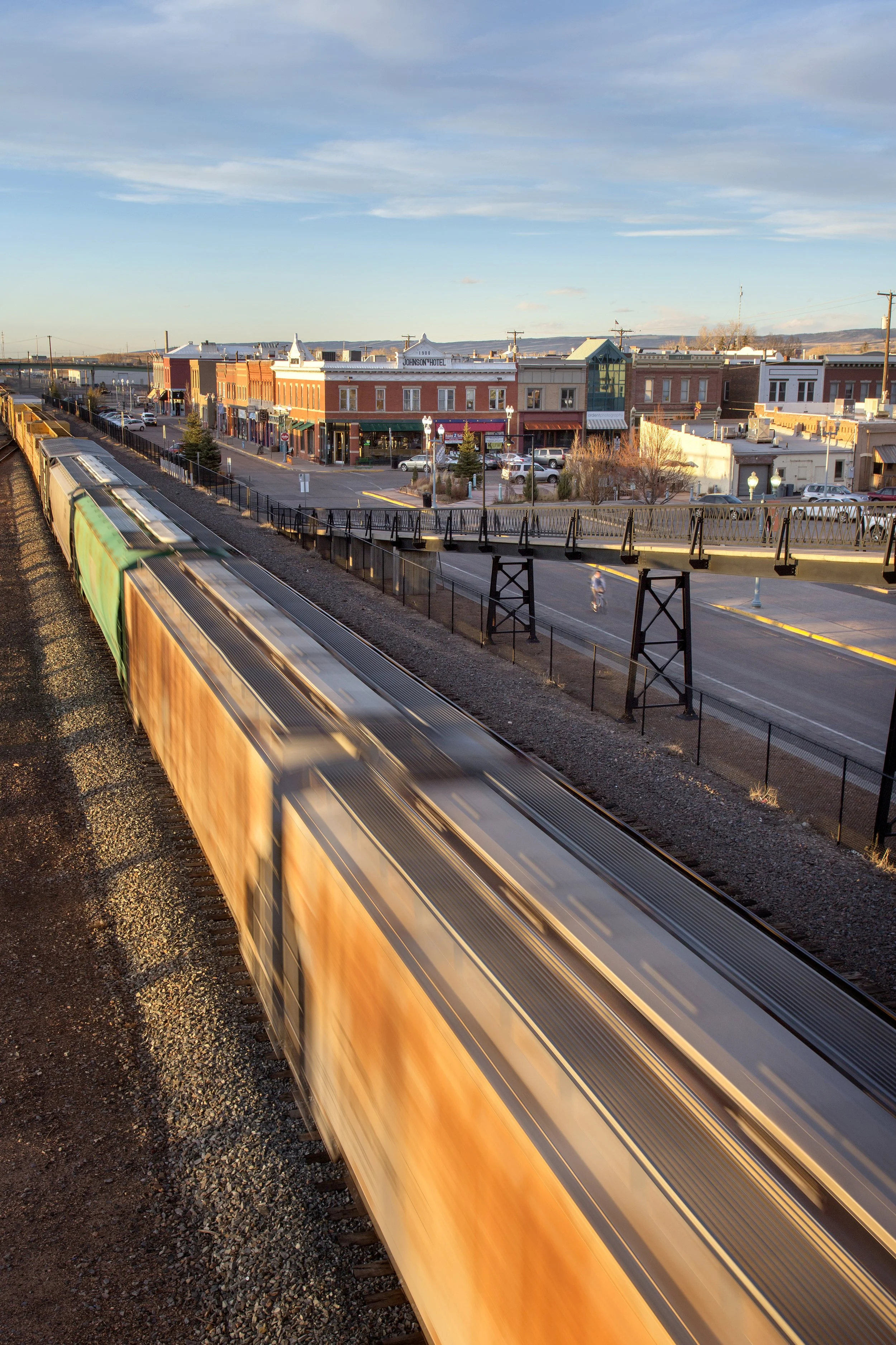 A moving train passes through a small town with brick buildings and storefronts, while a person rides a bicycle on the street and the sky is partly cloudy.