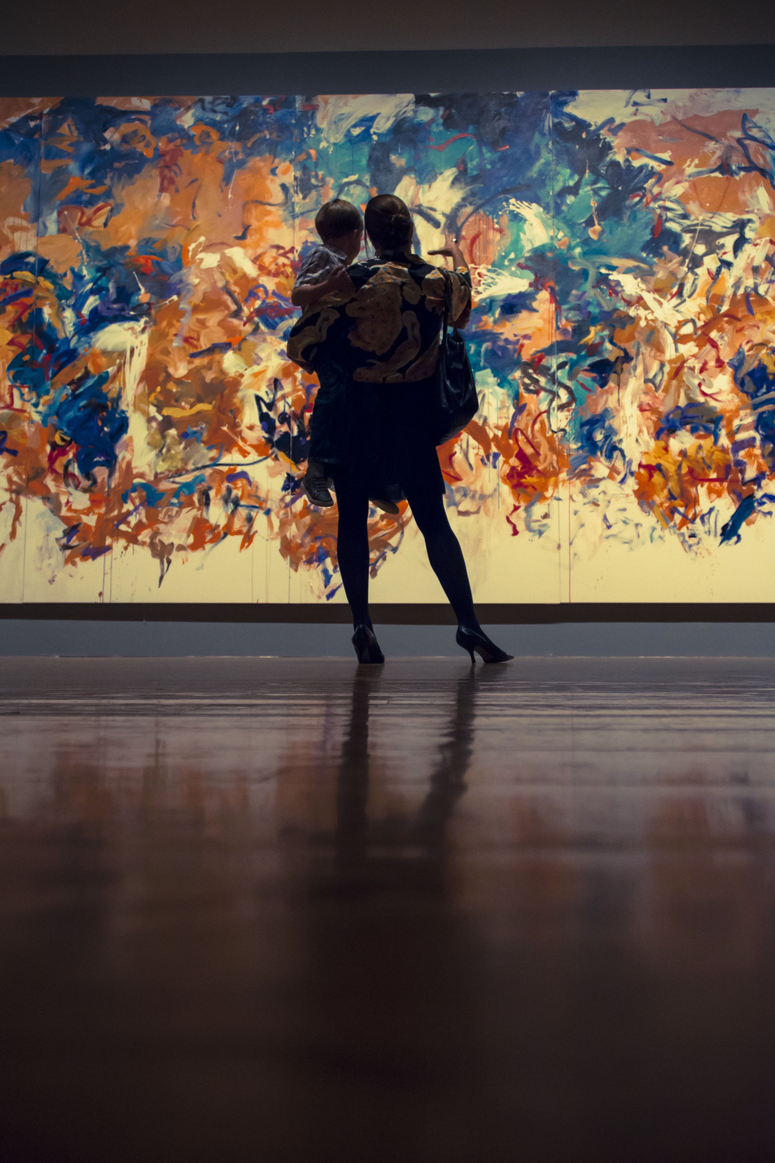 A woman in high heels and a patterned jacket holding a child, standing in front of an abstract colorful painting in a gallery.