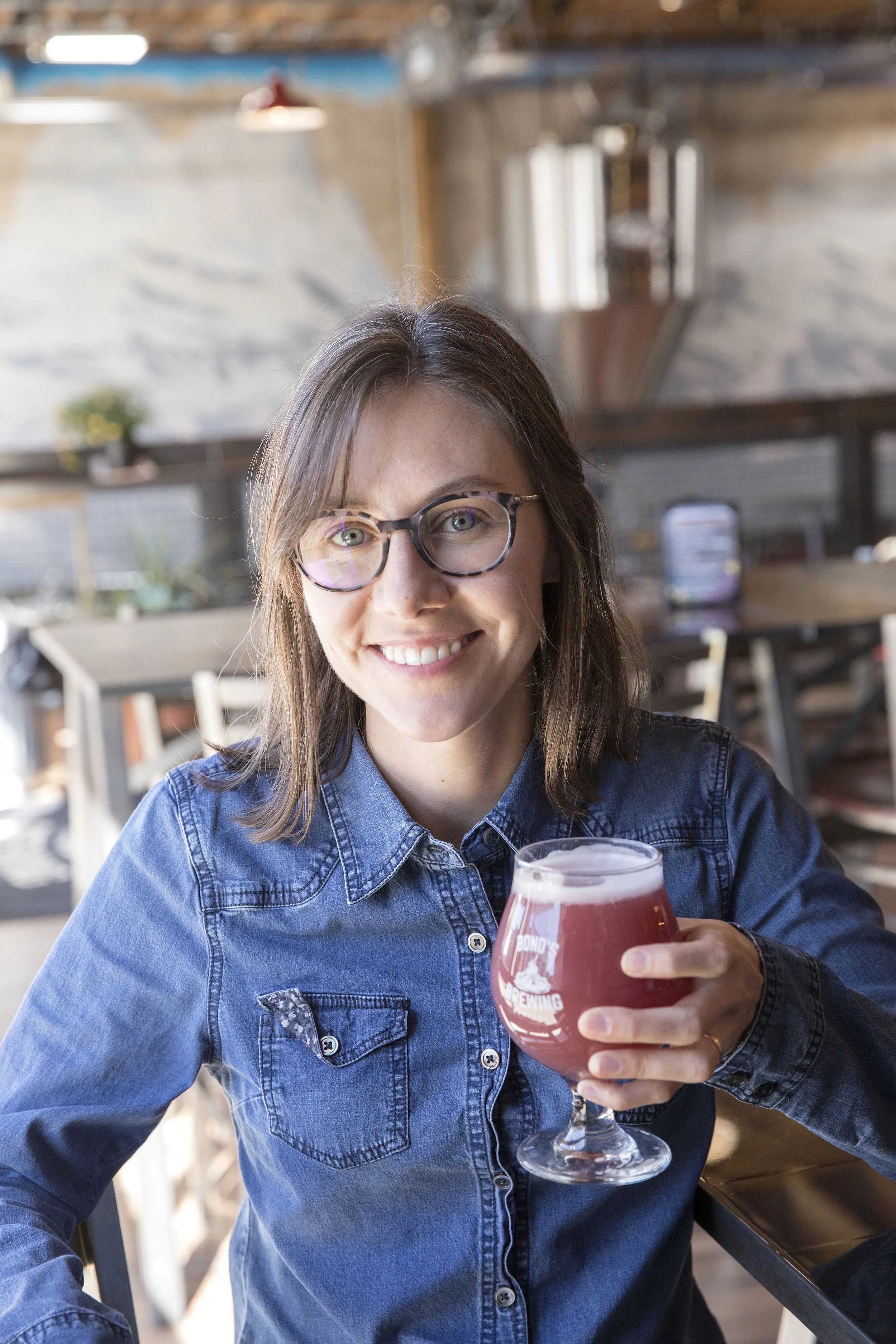 A woman with glasses and a denim shirt sitting at a table in a brewery, holding a glass of beer and smiling at the camera.