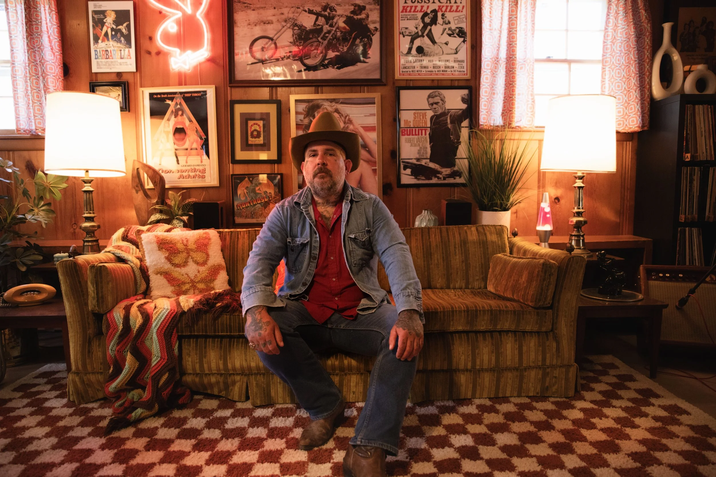 Country music artist Jeremy Pinnell wearing cowboy hat, red shirt, and denim jacket sitting on a vintage couch in a cozy, retro-style room with warm lighting, surrounded by framed posters and artwork on wood-paneled walls.
