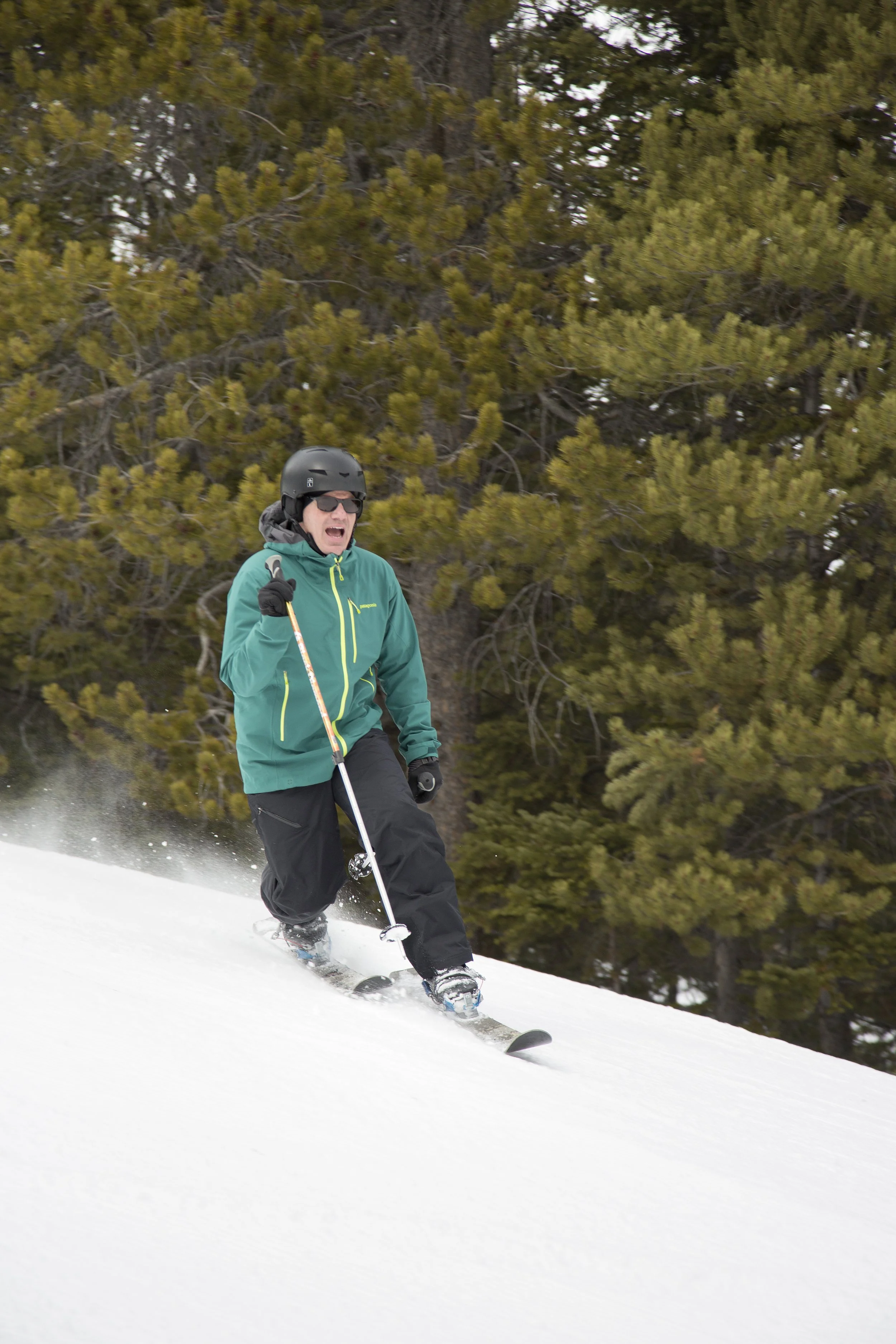 A person in a teal jacket, black helmet, and sunglasses skiing downhill on snow with trees in the background.