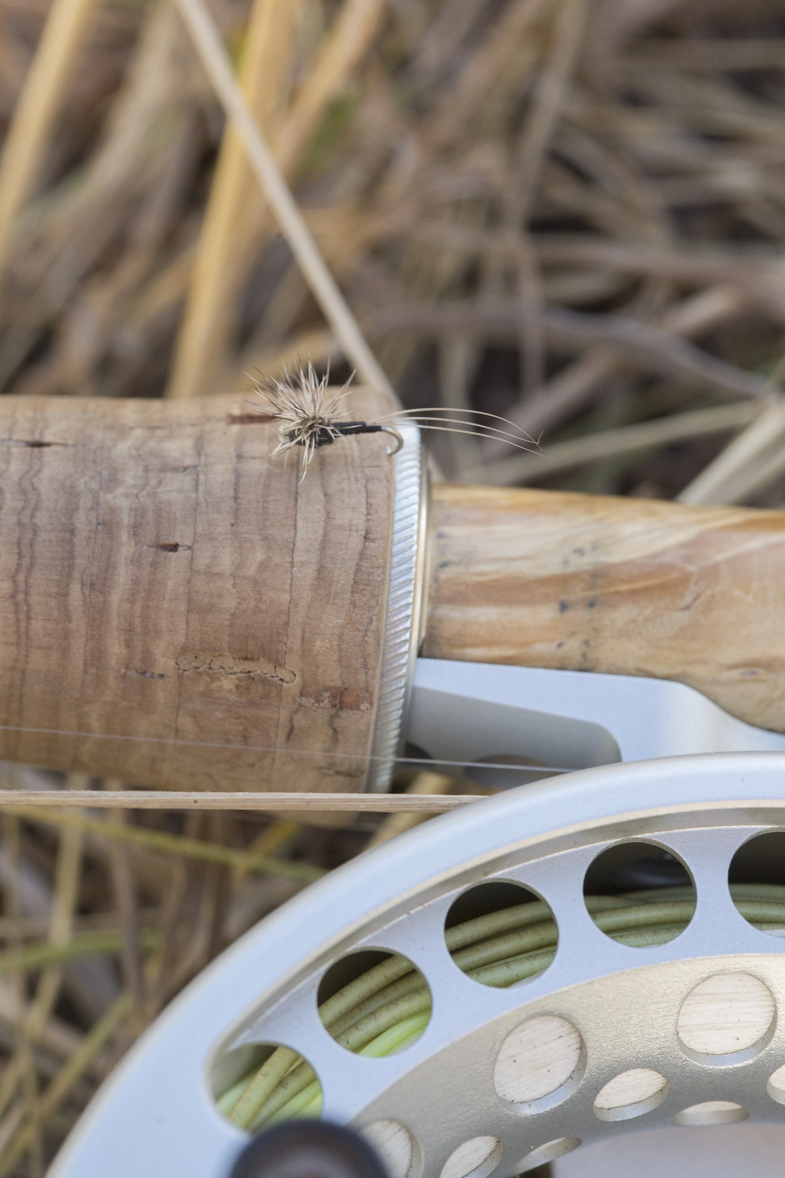 Close-up of a fishing fly on a wooden handle, with a metal handle and a fishing reel with green line visible in the background.