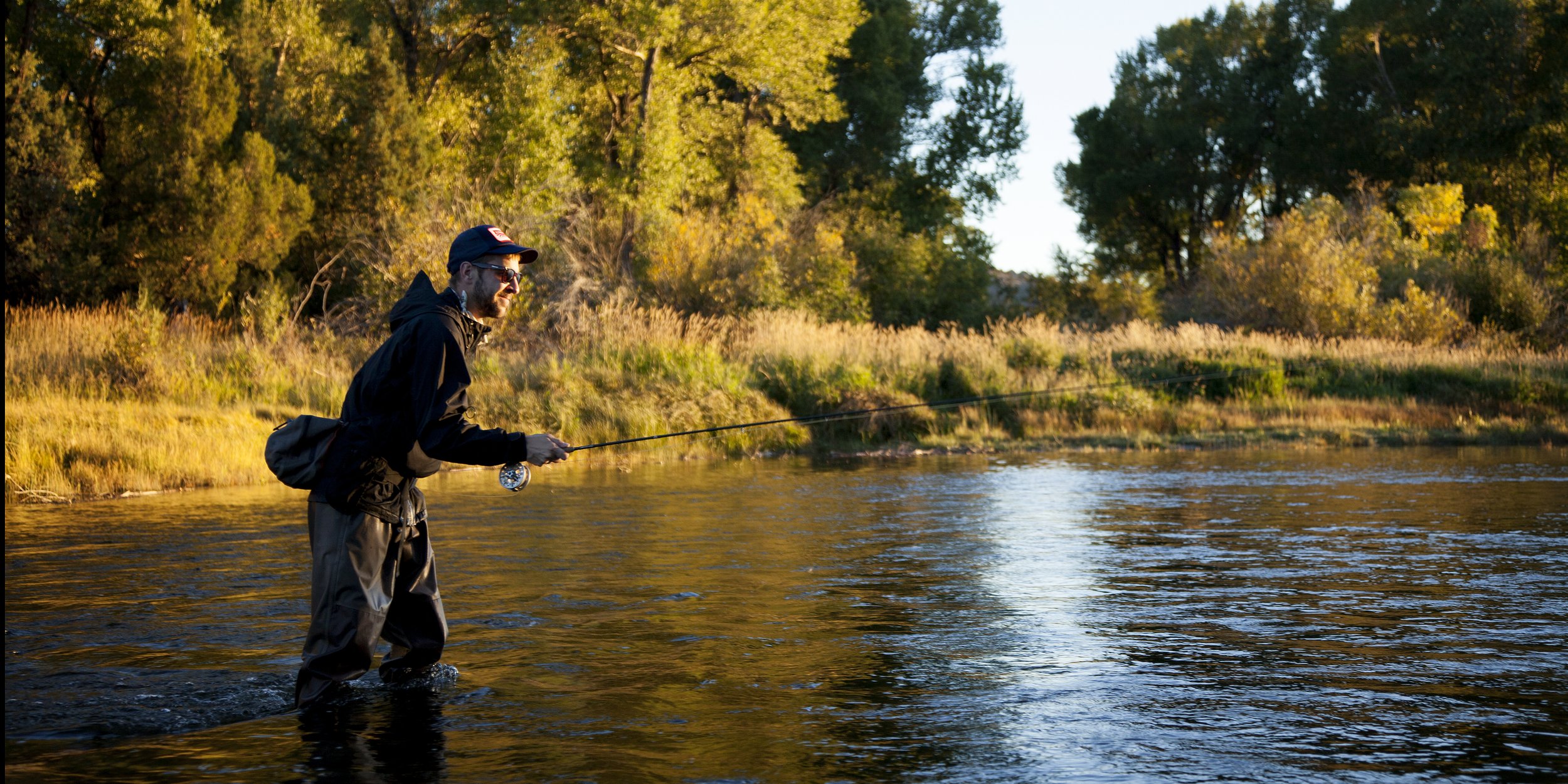 A man stands in a river, fishing with a rod, with trees and golden foliage in the background during sunset.