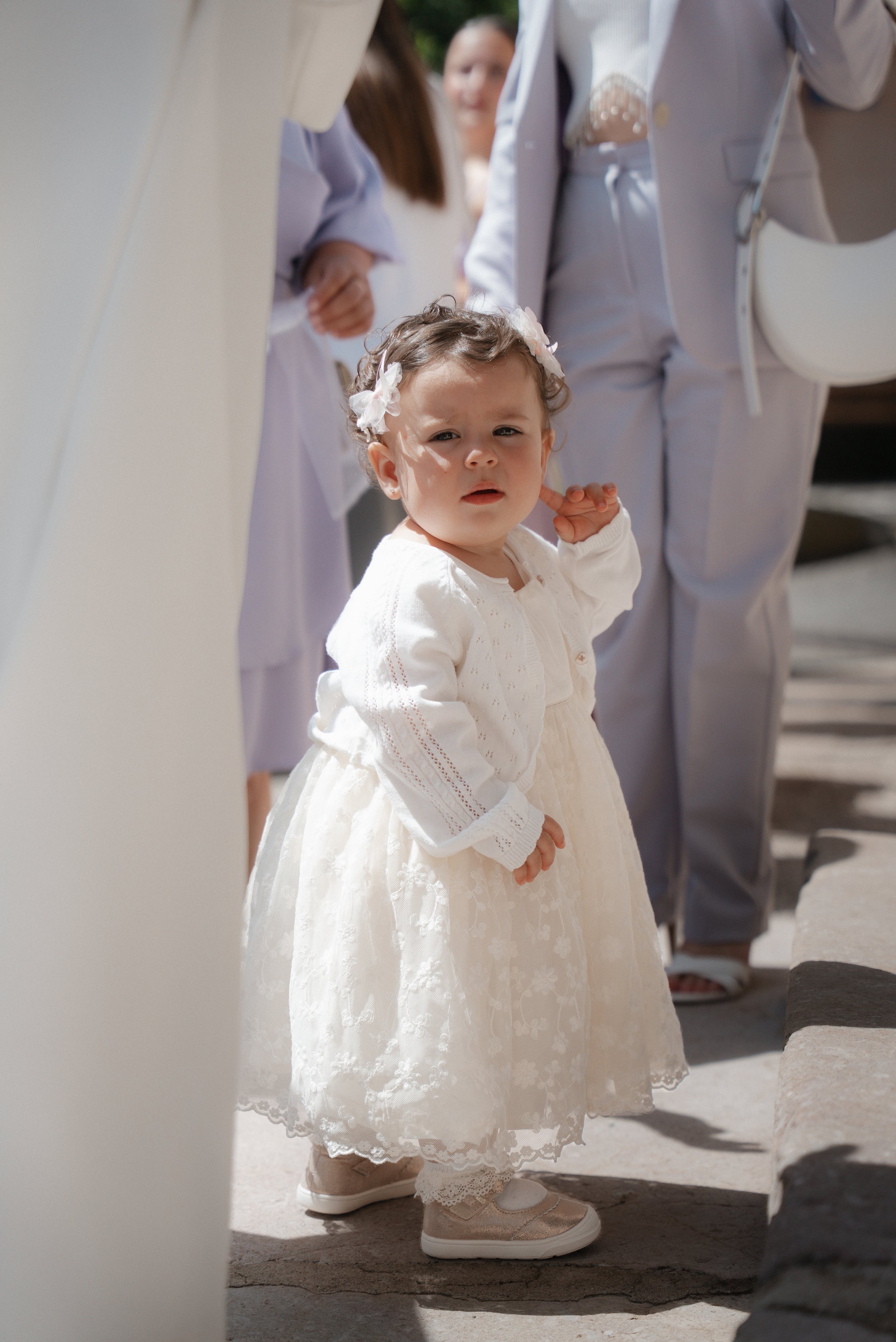 Une petite fille en robe blanche, portant une carde en laine blanche et des chaussures roses, se tient parmi un groupe de personnes lors d'une cérémonie en extérieur.