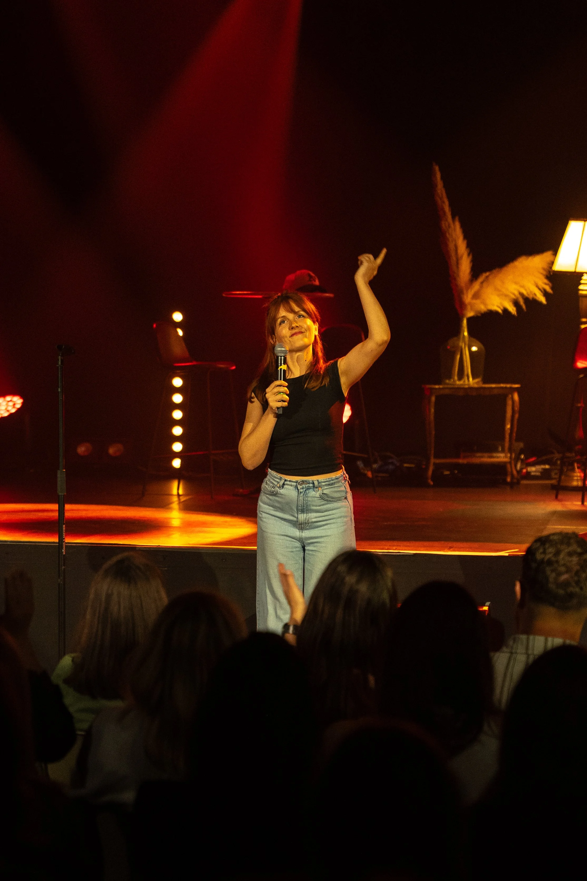 Une jeune femme avec des cheveux roux, portant un haut noir et un jean clair, tient un micro dans la main gauche et lève la main droite lors d'une performance sur scène devant un public, avec un décor chaleureux comprenant des lampes et des plantes.
