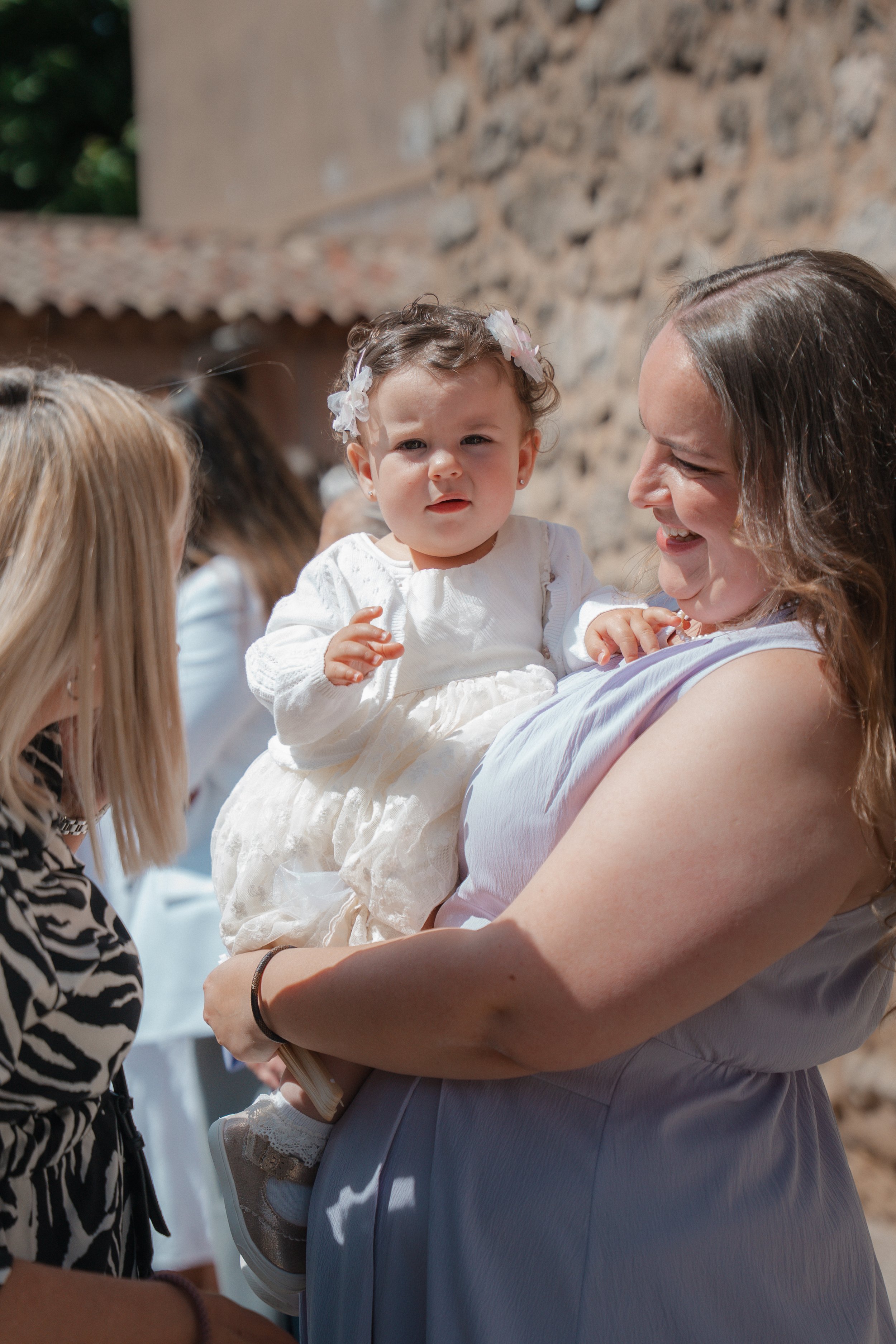 Une femme porte un bébé lors d'une célébration en extérieur, avec un mur en pierre en arrière-plan.