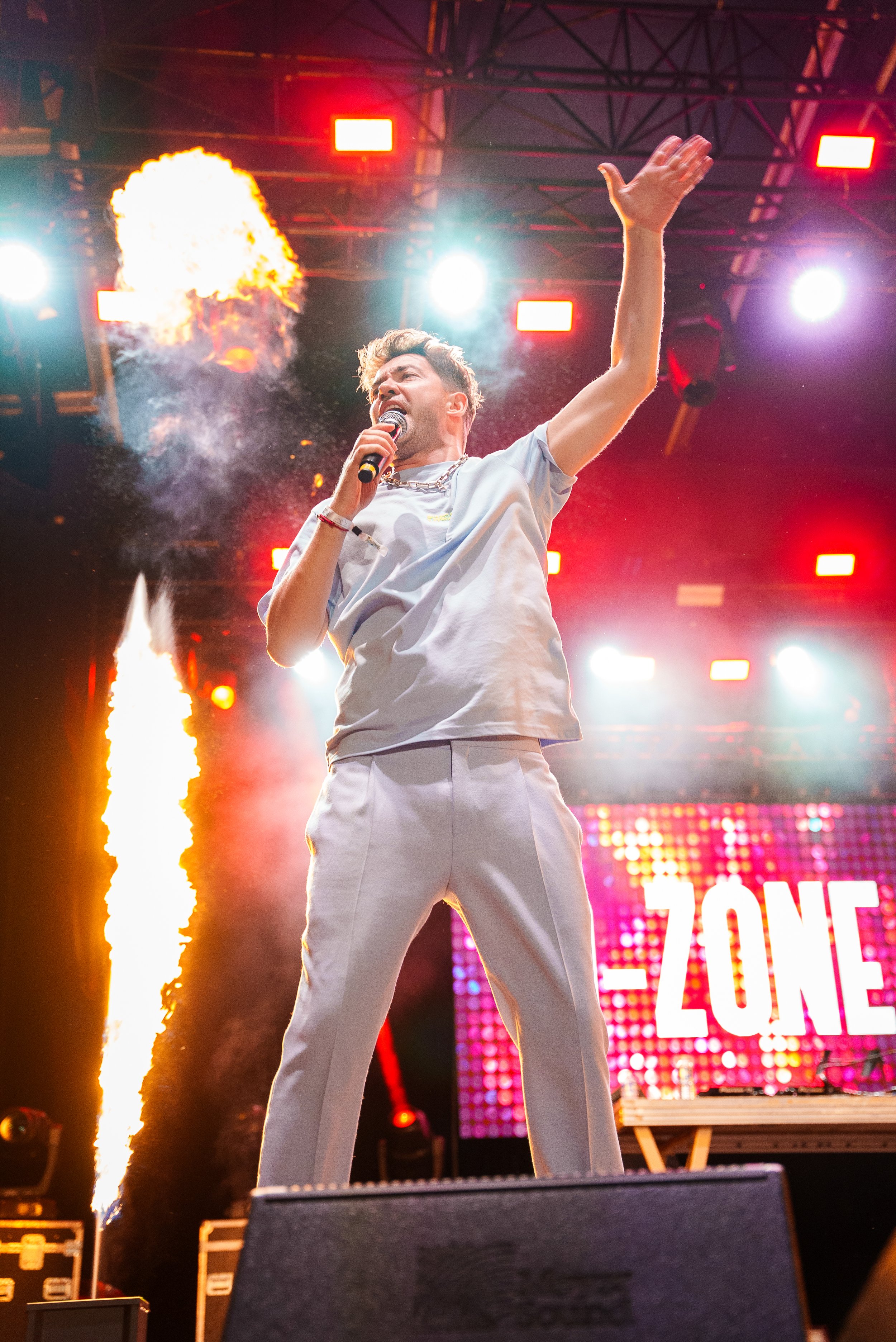 Un chanteur sur scène en train de performer, avec des effets pyrotechniques et un éclairage coloré, devant un écran affichant le mot "ZONE".