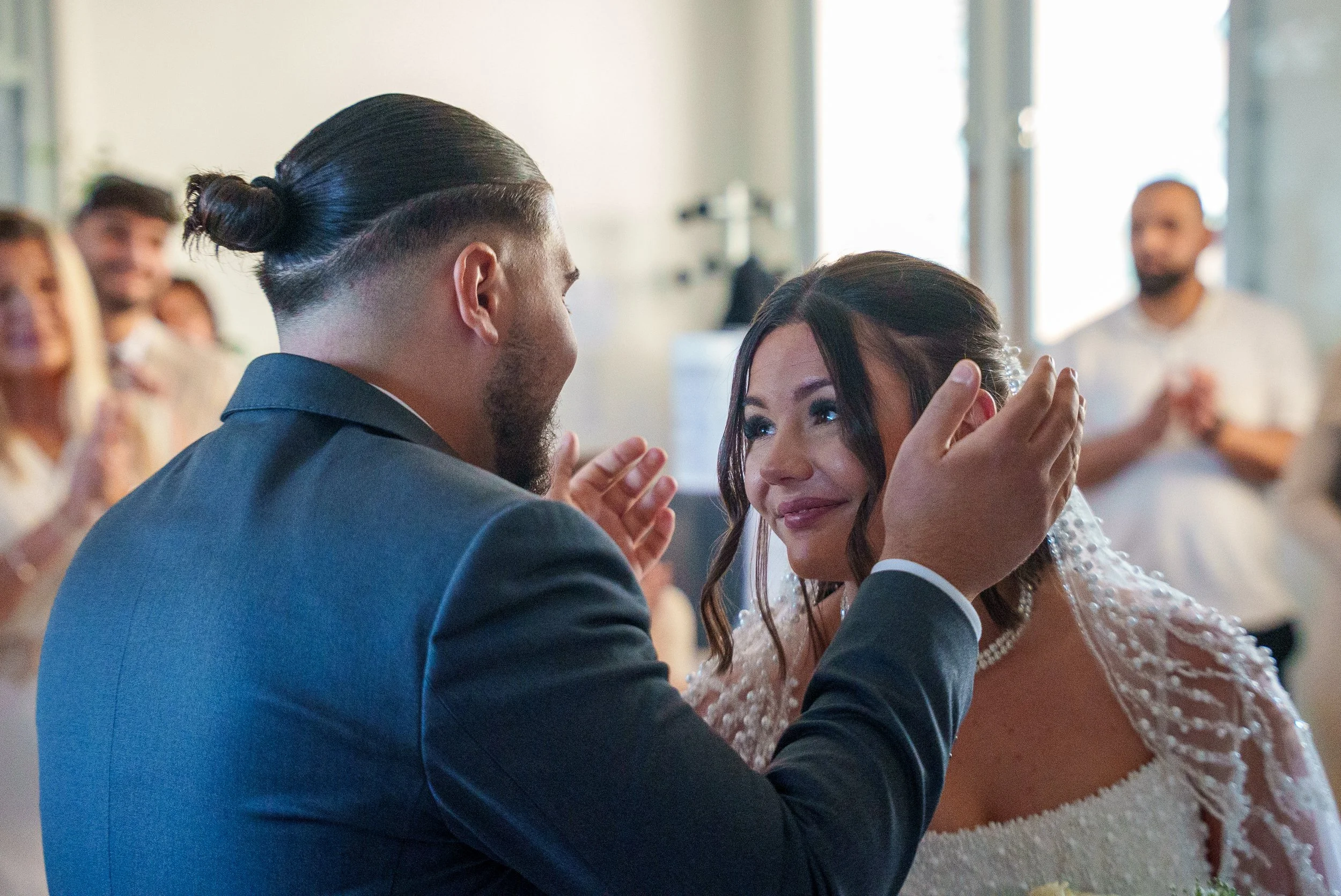 Un homme et une femme lors de leur mariage, l'homme lui tient le visage et ils se regardent avec émotion, entourés d'amis et de famille qui sourient