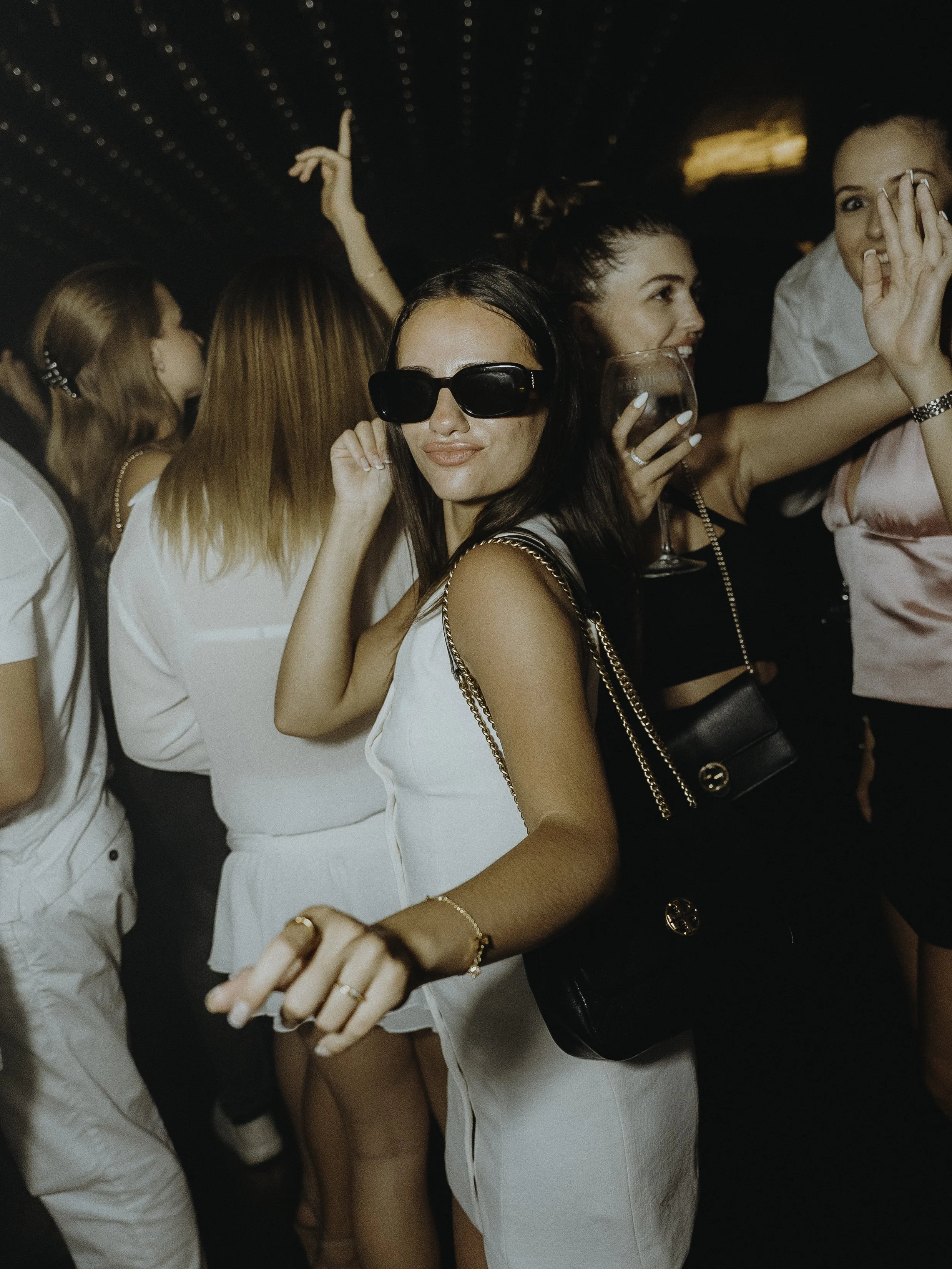 Groupe de jeunes femmes lors d'une fête ou soirée, portant des vêtements élégants et des sacs à main, dans une ambiance festive avec des lumières de fête, en train de danser et de socialiser.