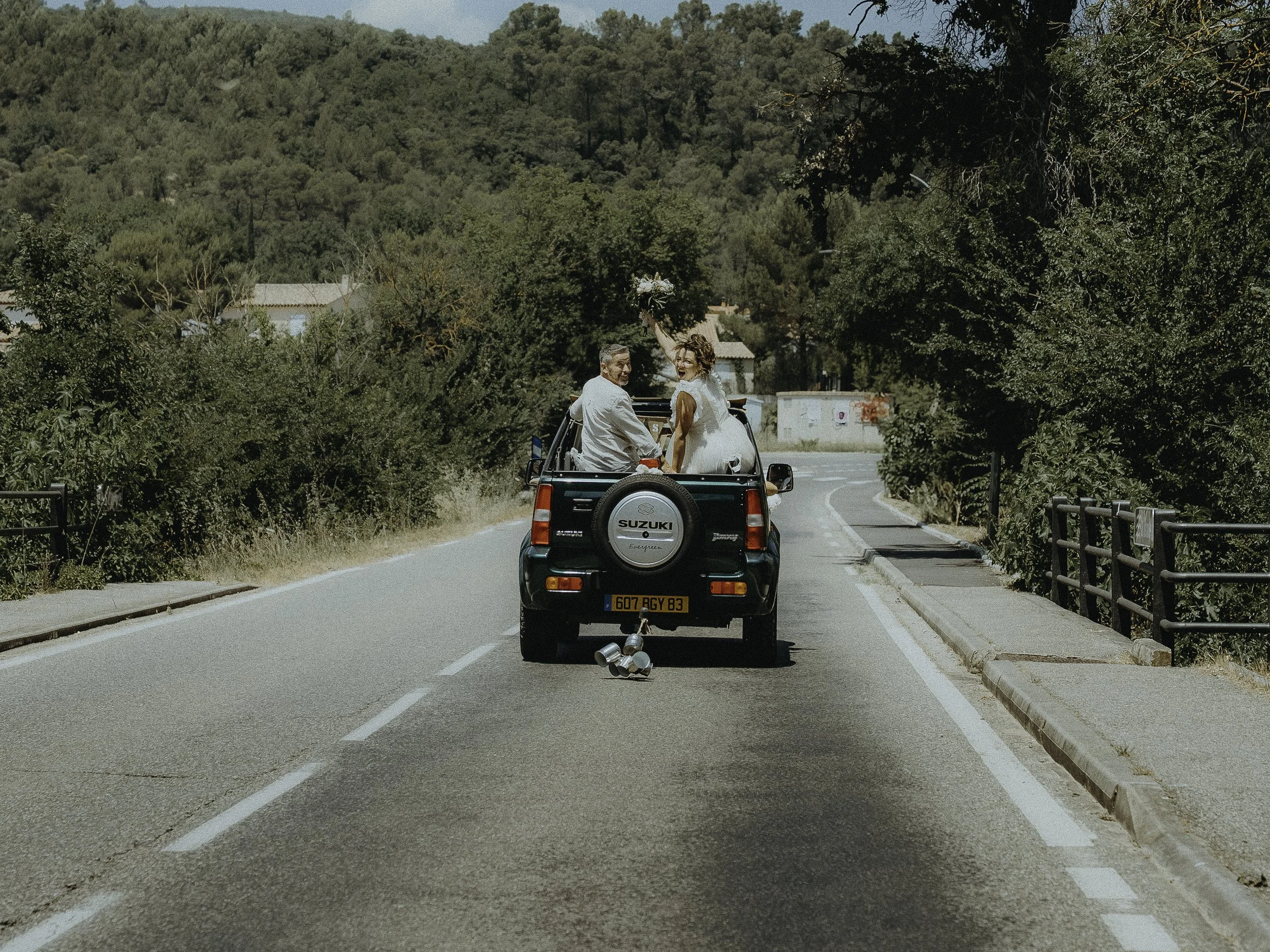 Deux personnes assises à l'arrière d'une petite voiture jaune, roulant sur une route de campagne entourée d'arbres, avec une femme levant un bouquet de fleurs et un homme à côté d'elle.