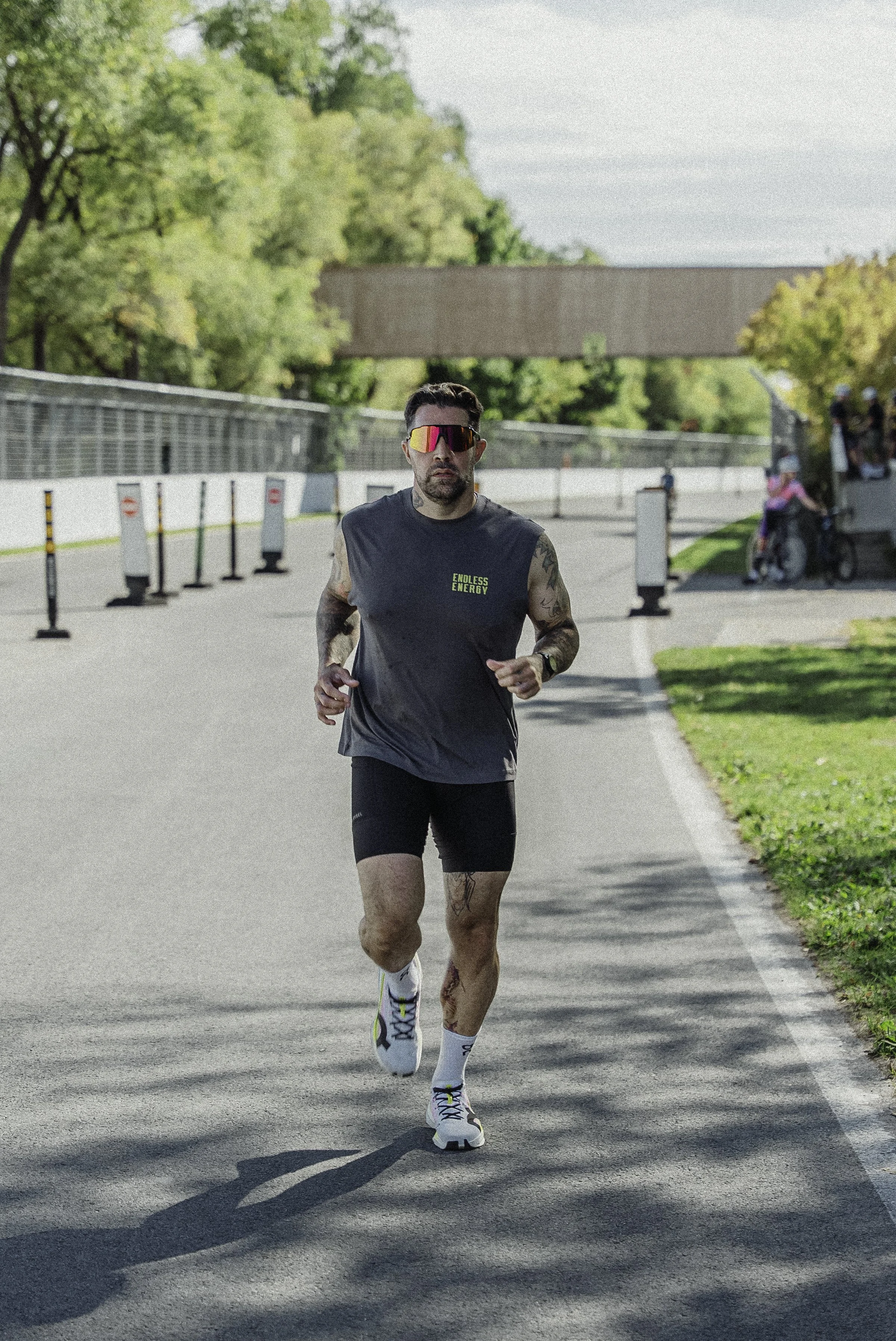 Homme courant sur une route en extérieur, portant un t-shirt sans manches, un short de course, des lunettes de soleil rouges et une montre. Arbres et autres coureurs en arrière-plan.