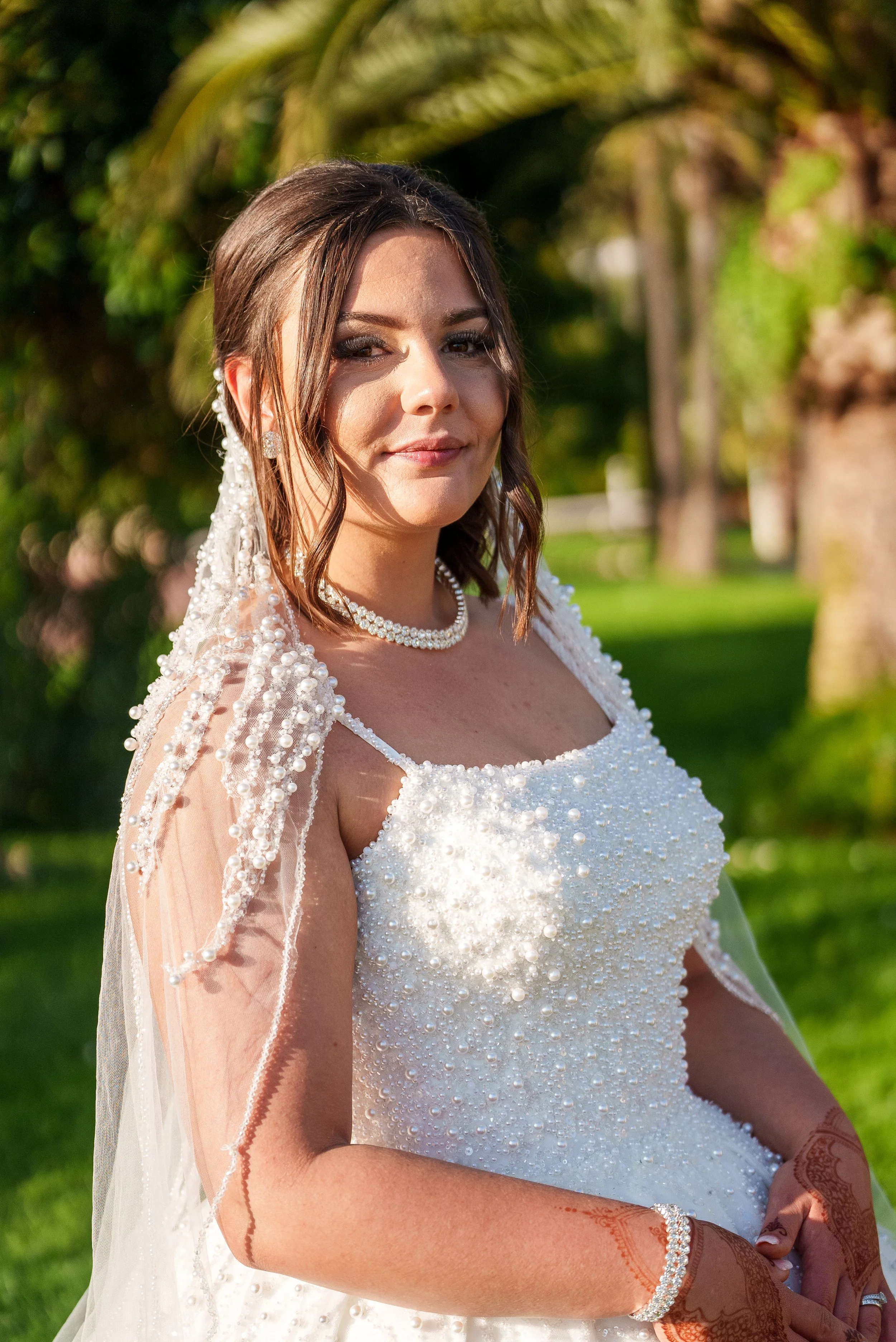 Jeune femme souriante en robe de mariée, portant des perles, avec des tatouages sur les bras, portant un voile et des bijoux, en extérieur avec des arbres en arrière-plan.