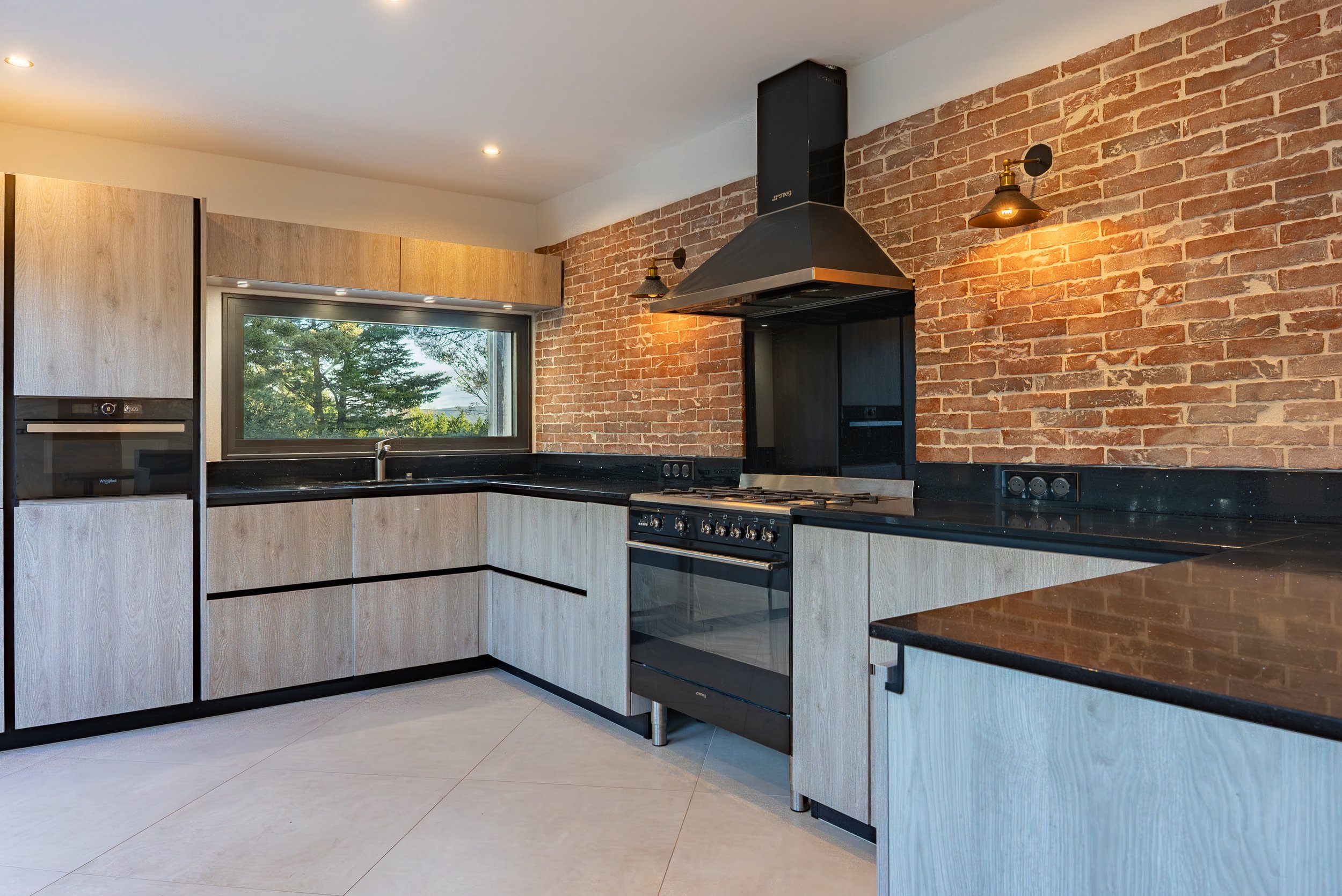 Cuisine moderne avec murs en briques visibles, comptoirs noirs en granite, fenêtres laissant entrer la lumière naturelle, et mobilier en bois clair. Éléments de cuisine intégrés, notamment une cheminée, et éclairage mural.