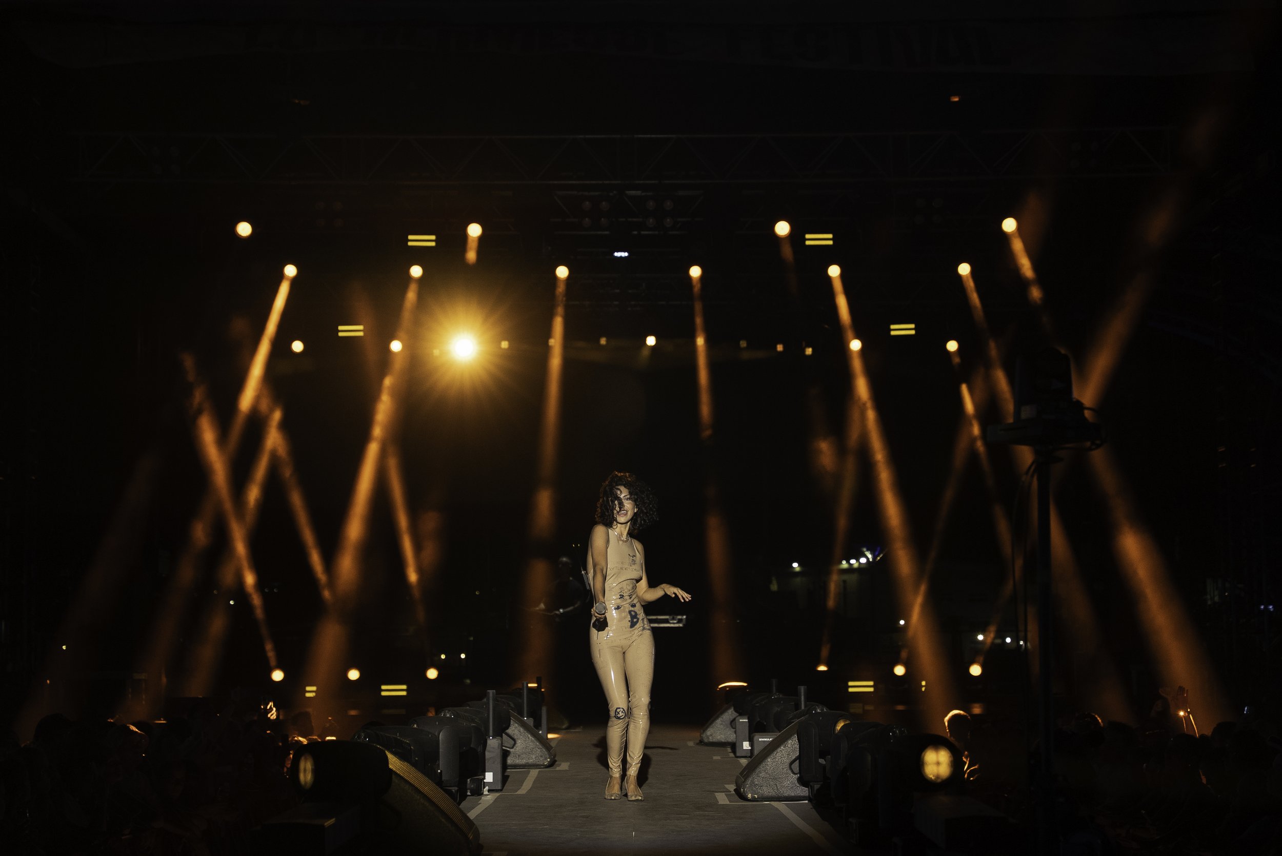Une chanteuse debout sur scène lors d'un concert, entourée de lumières orange et jaunes, avec un fond sombre.