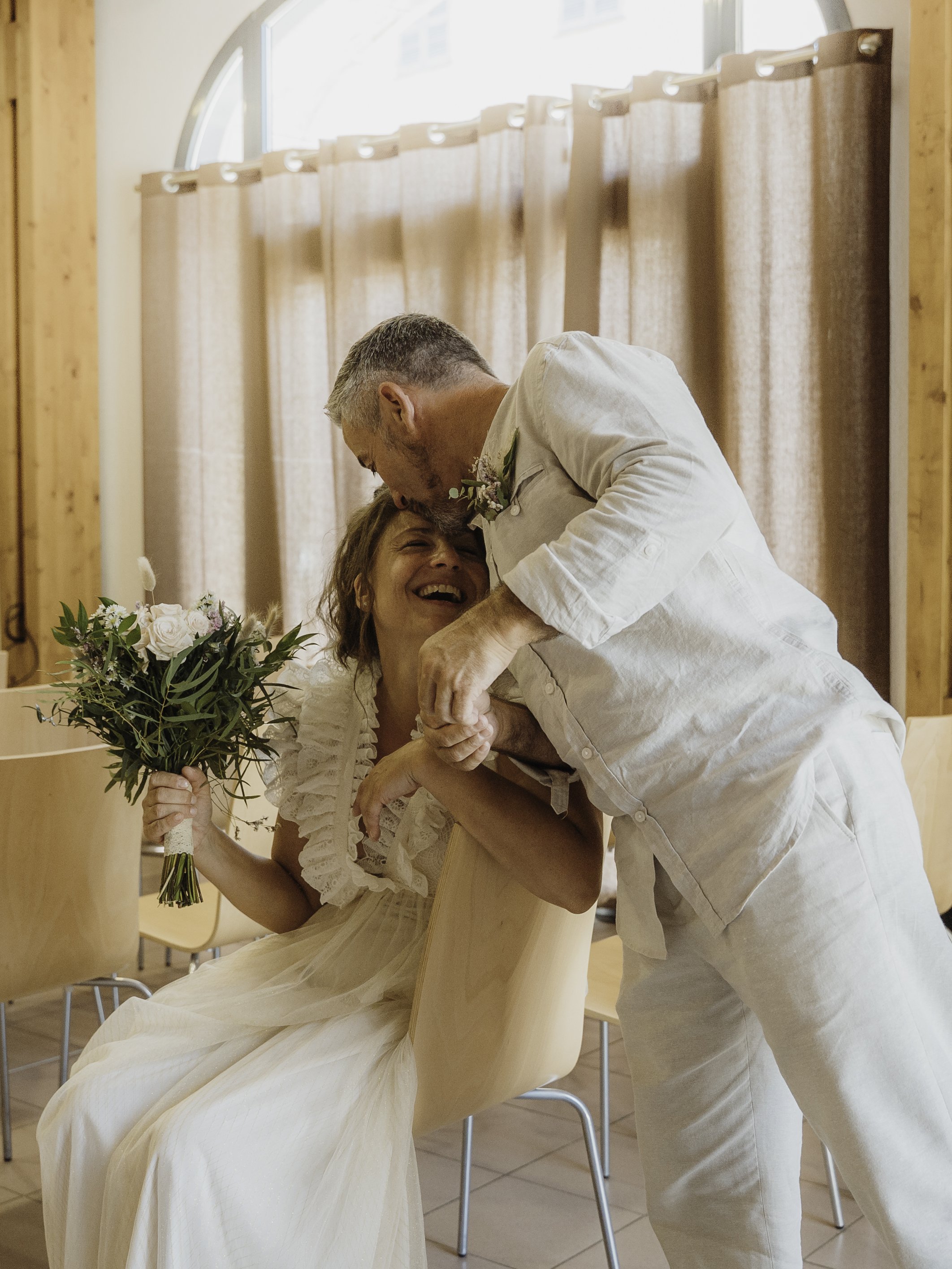 Un homme et une femme partagent un moment de joie et d'amour lors d'une cérémonie. La femme tient un bouquet de fleurs et sourit tandis que l'homme l'embrasse sur le front.
