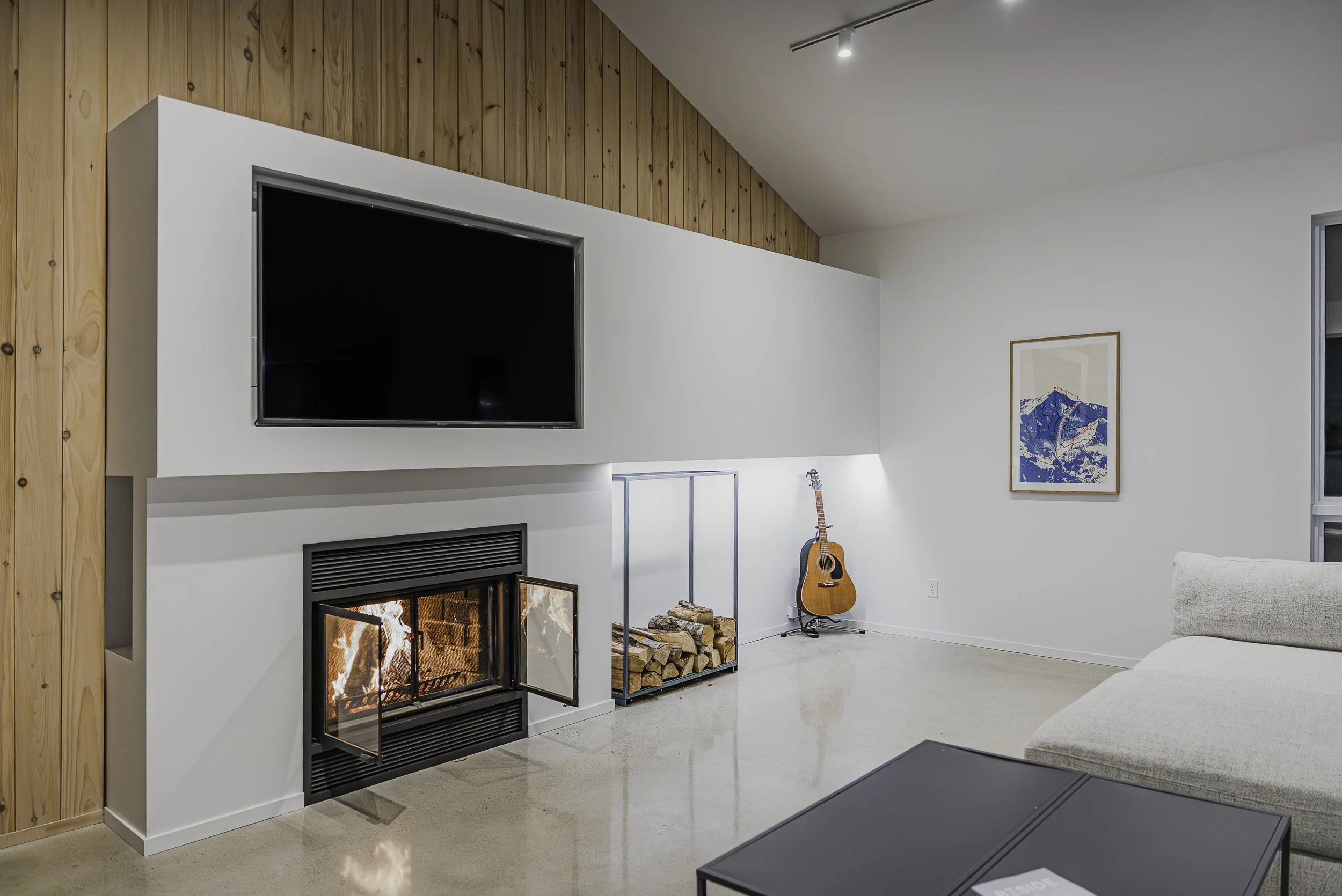Salon moderne avec cheminée allumée, grande télévision à écran plat, guitare acoustique, architecture épurée avec murs blancs et boiserie en haut, tableau de montagnes, canapé blanc et table noire.