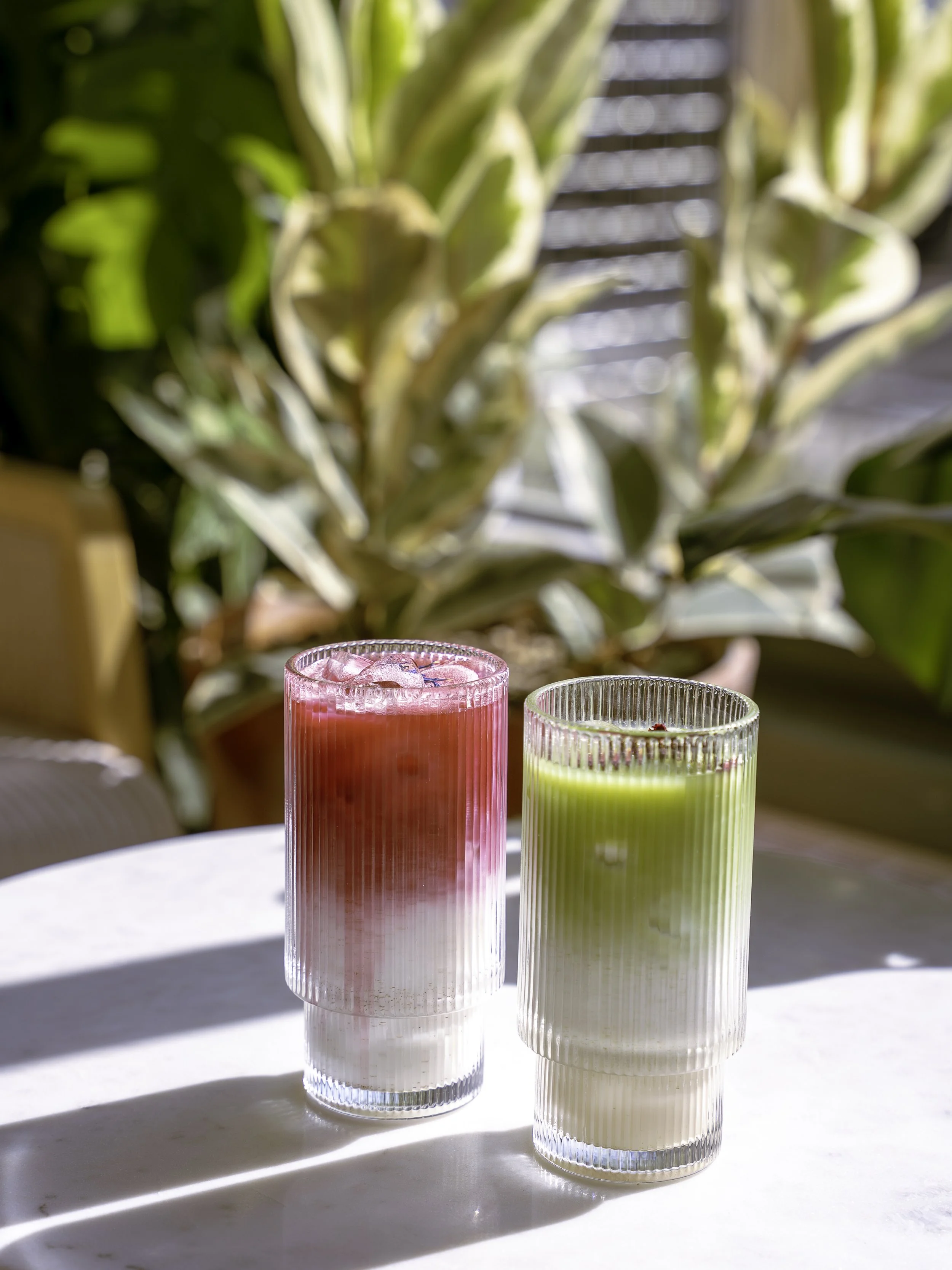 Deux verres en verre transparent avec des boissons colorées, l'une rose et l'autre vert pâle, placés sur une table blanche avec des ombres de lumière naturelle, avec une plante verte en arrière-plan
