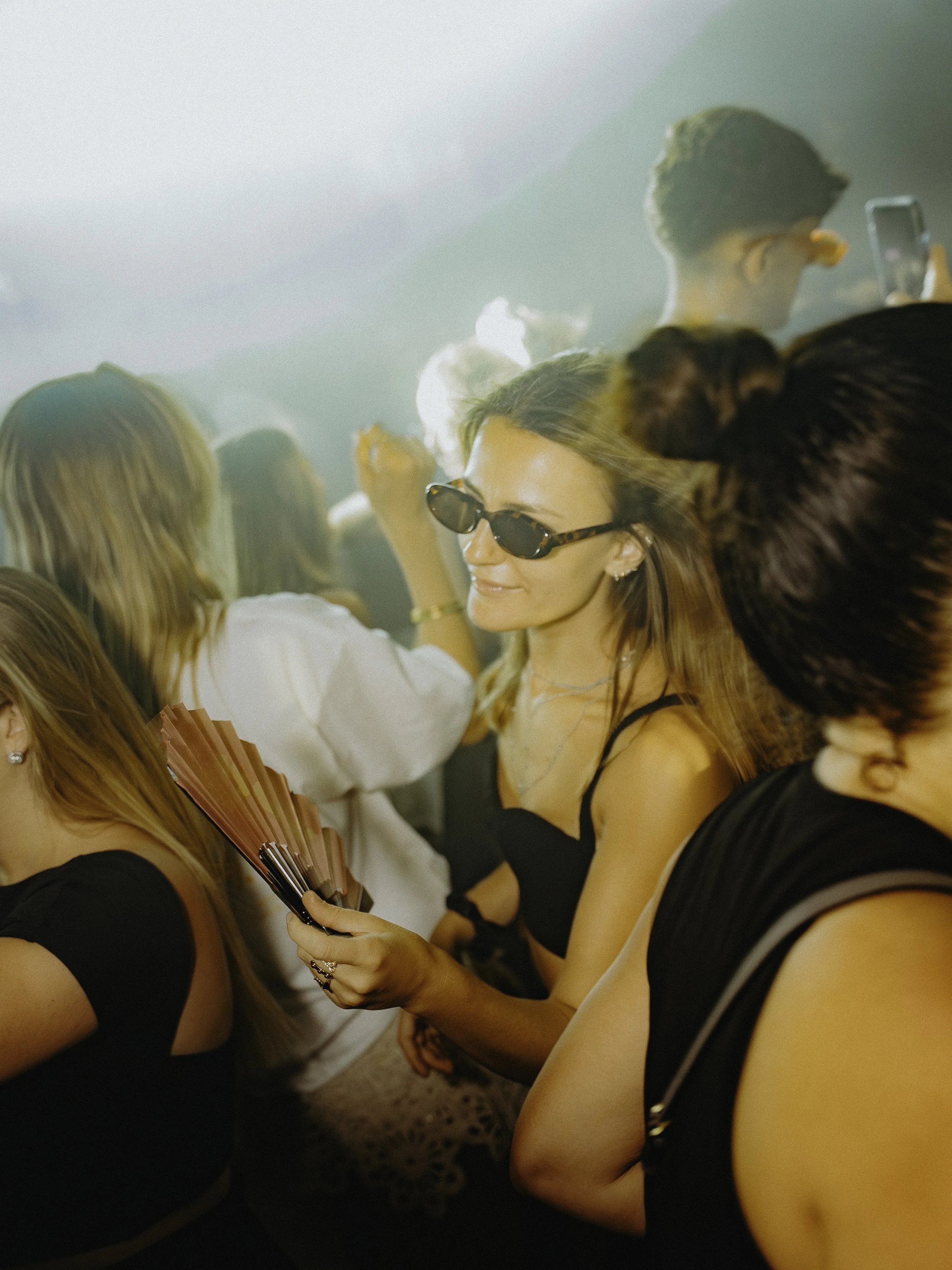 Une femme portant des lunettes de soleil tient un éventail lors d'une fête ou d'un concert, entourée d'autres personnes dans une ambiance lumineuse et festive.