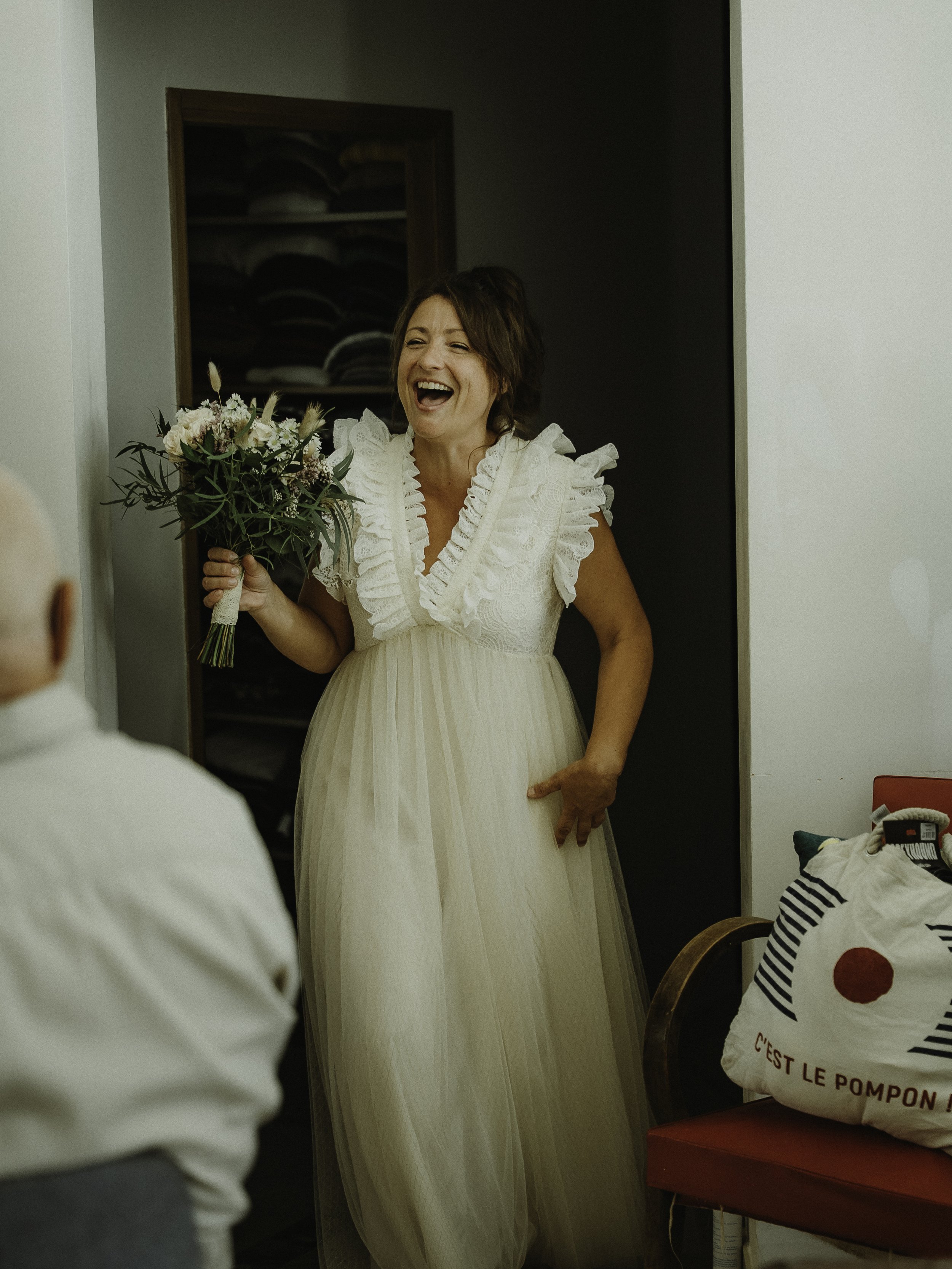 Une femme souriante en robe blanche avec un bouquet de fleurs, probablement une mariée, dans une pièce intérieure.
