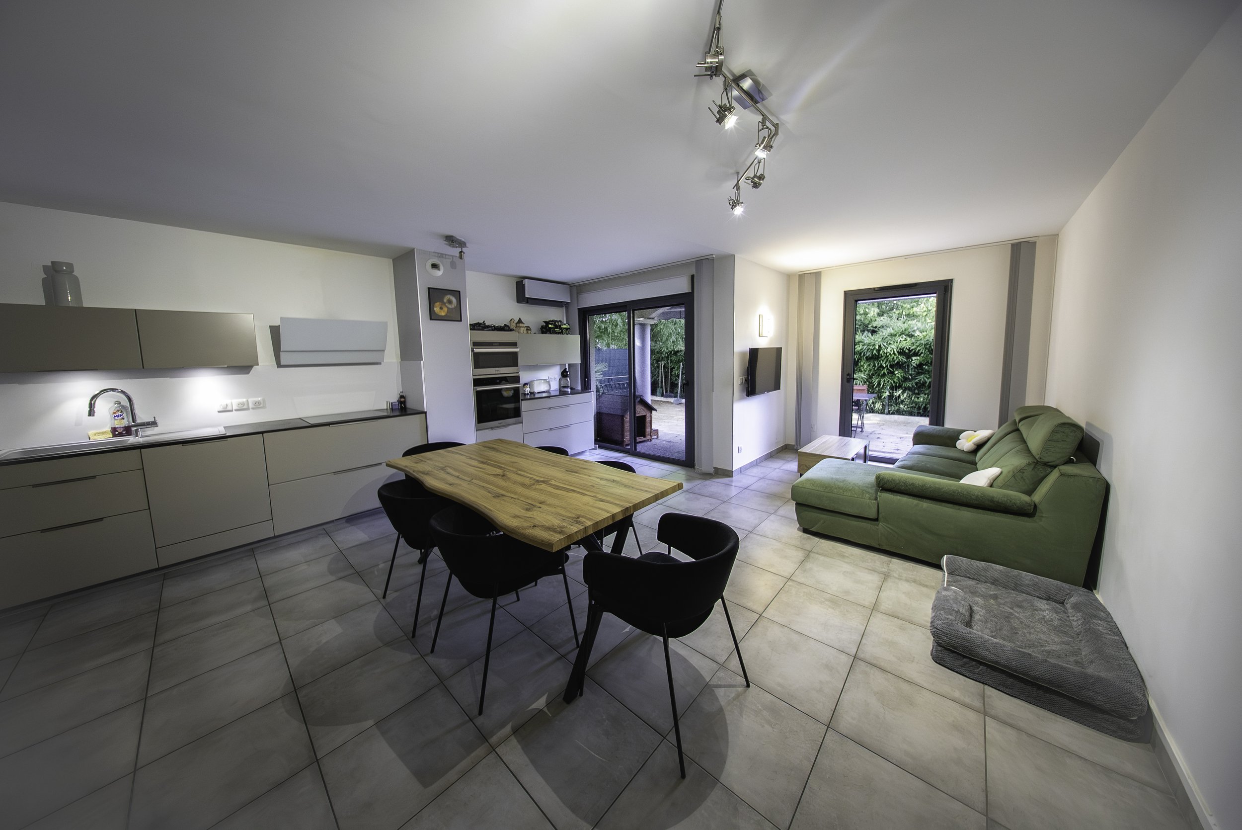Salle de séjour ouverte avec cuisine moderne, table en bois avec six chaises noires, canapé vert, porte-fenêtre donnant sur un jardin, et un mur avec télévision.