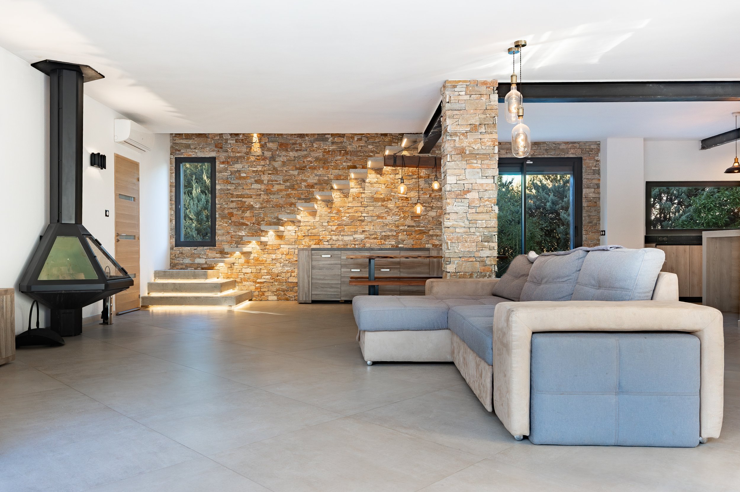Salon moderne avec canapé gris, mur en pierre apparente, escaliers lumineux, cheminée en coin, grande fenêtre et éclairage suspendu.