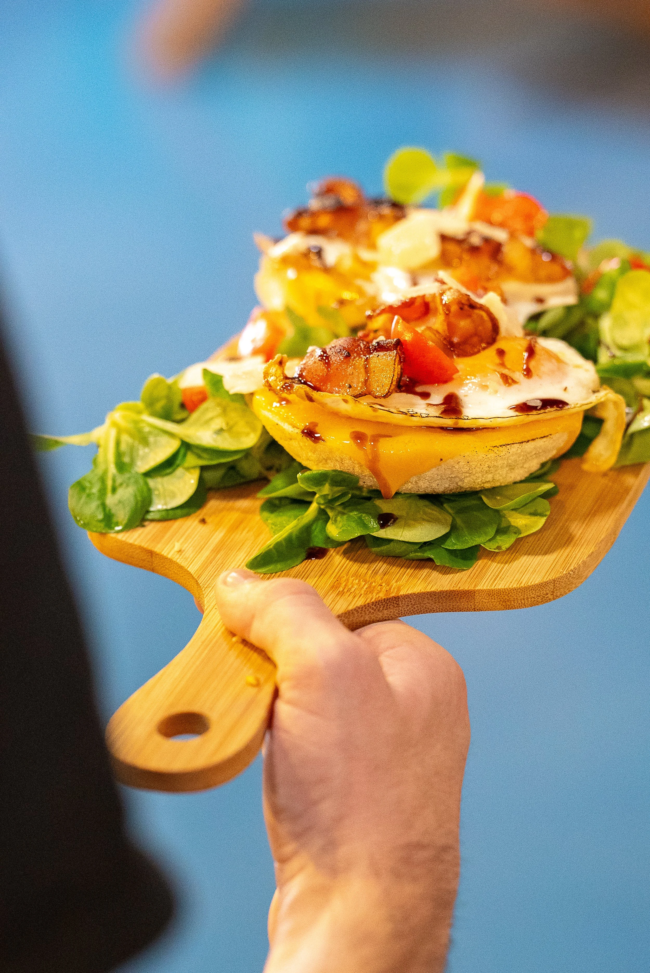 Une planche en bois avec deux muffins contenant des œufs frits, garnis de bacon, tomates et fromage, sur un lit de jeunes pousses de salade.
