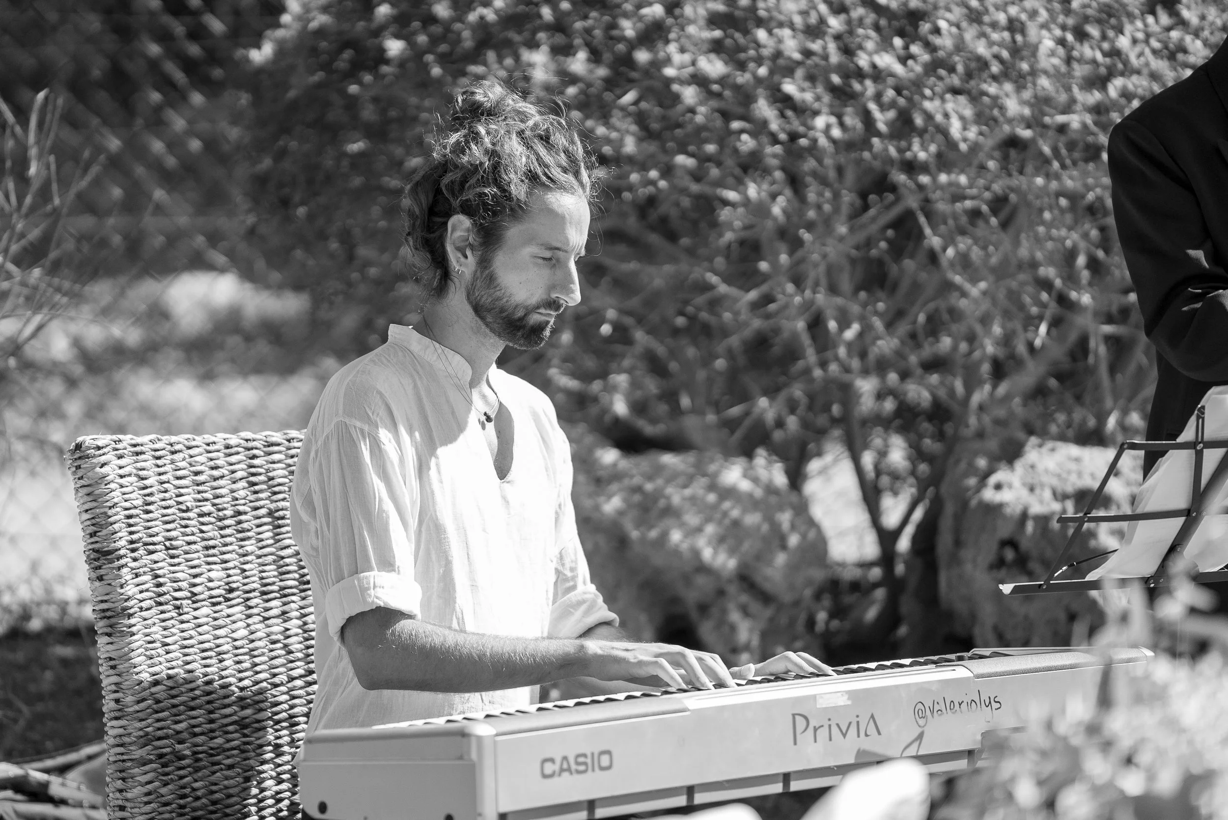 Un homme jouant du piano à l'extérieur, assis sur une chaise en osier, avec des arbres en arrière-plan, en noir et blanc.