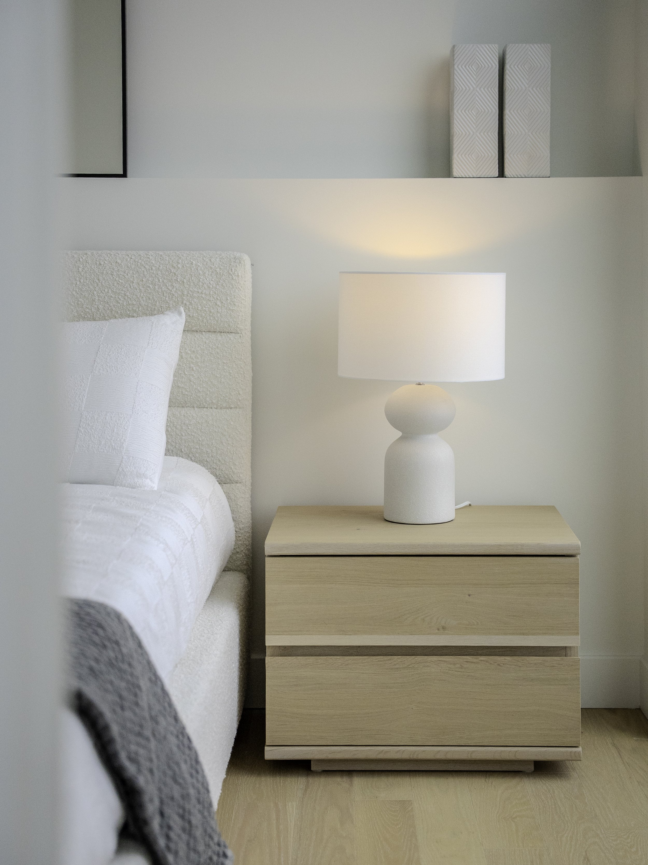 Chevet en bois clair avec lampe blanche à base ronde, dans une chambre avec lit à headboard en tissu crème et oreiller blanc, murs blancs et deux livres empilés en haut.