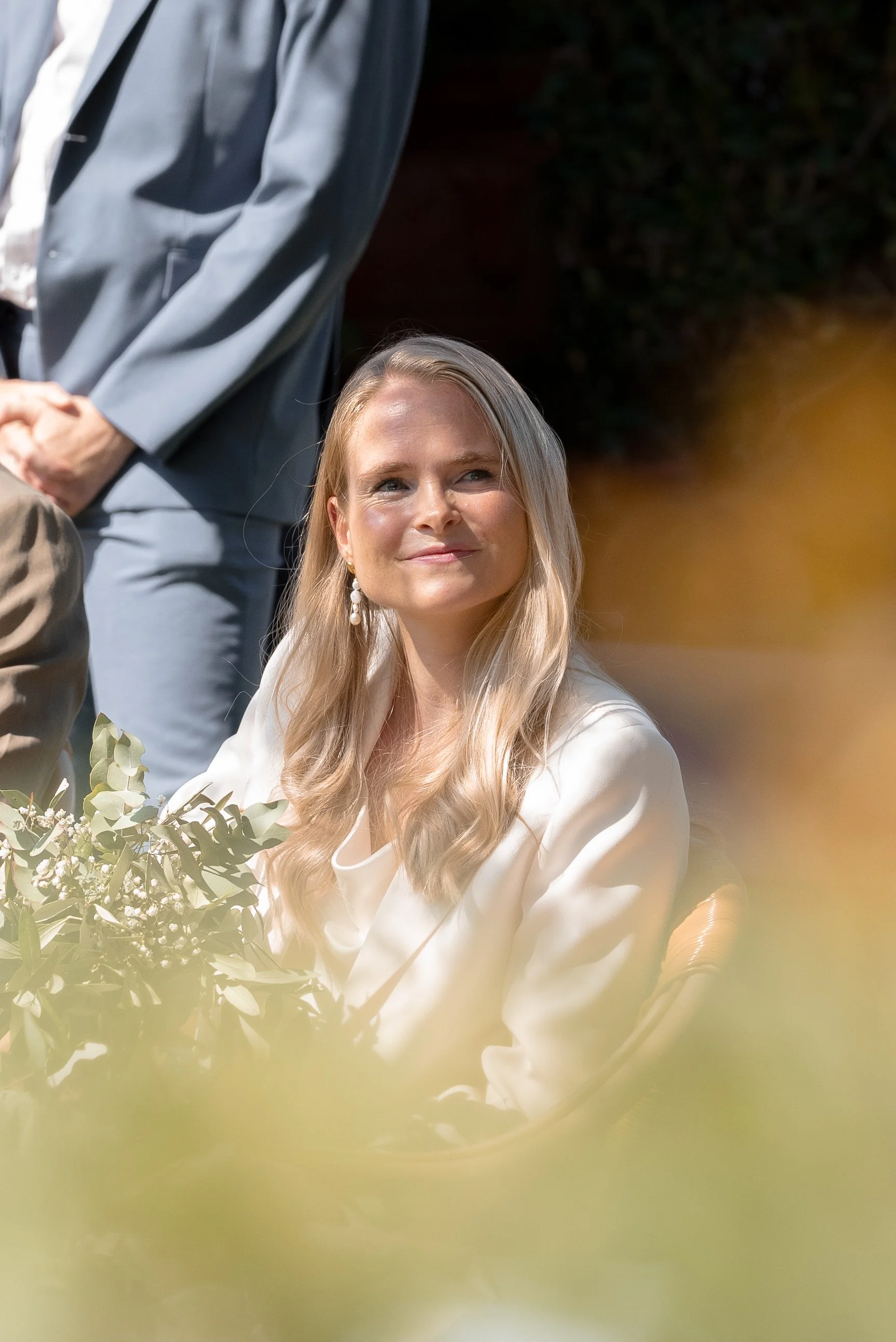 Une femme blonde souriante assise à une table, tenant un bouquet de fleurs, lors d'un événement en plein air.